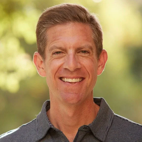 A smiling man with short, light brown hair wearing a collared shirt outdoors with blurred greenery in the background.