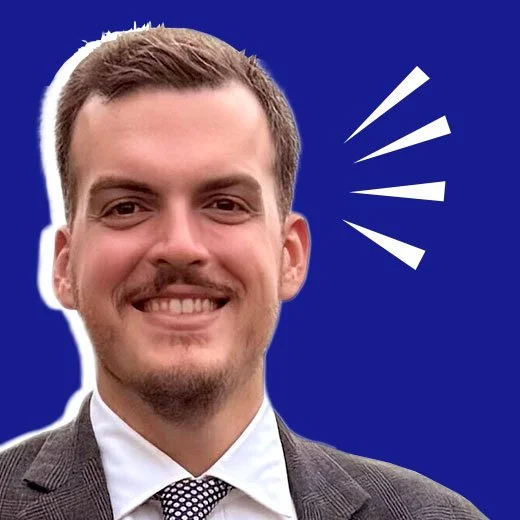 A smiling young man with short hair and a beard, wearing a suit and tie, with blue background and white graphic lines.