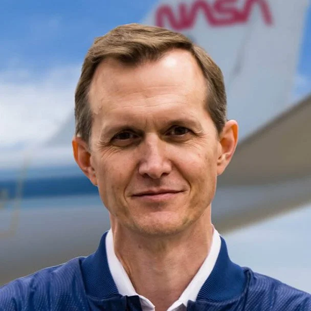 Man in blue jacket standing in front of airplane with NASA logo in background.