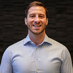 A smiling man in a light blue button-up shirt standing against a dark textured background.