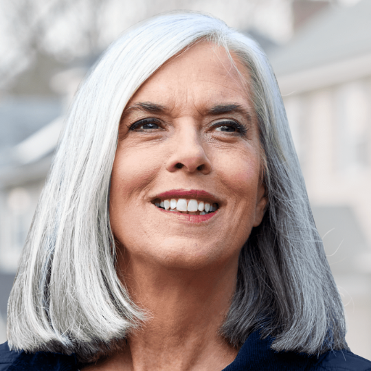 Close-up of a smiling older woman with long gray hair, outdoors on a bright day.