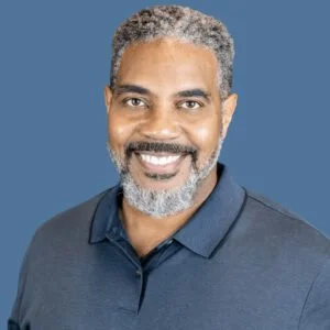 A smiling man with gray hair and a beard, wearing a dark blue collared shirt, against a blue background.
