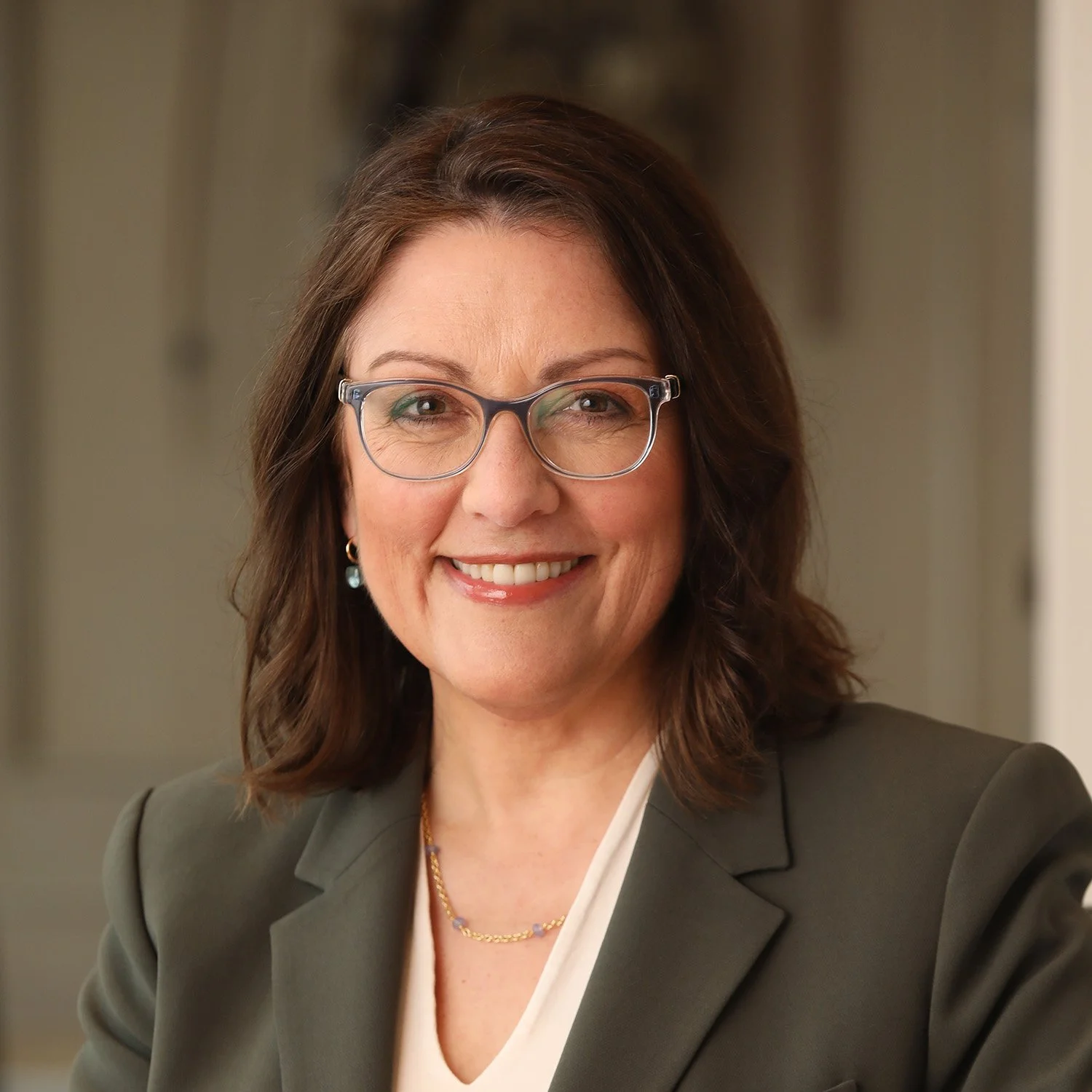 A woman with shoulder-length brown hair, wearing glasses, earrings, a necklace, a white blouse, and a brown blazer, smiling at the camera.