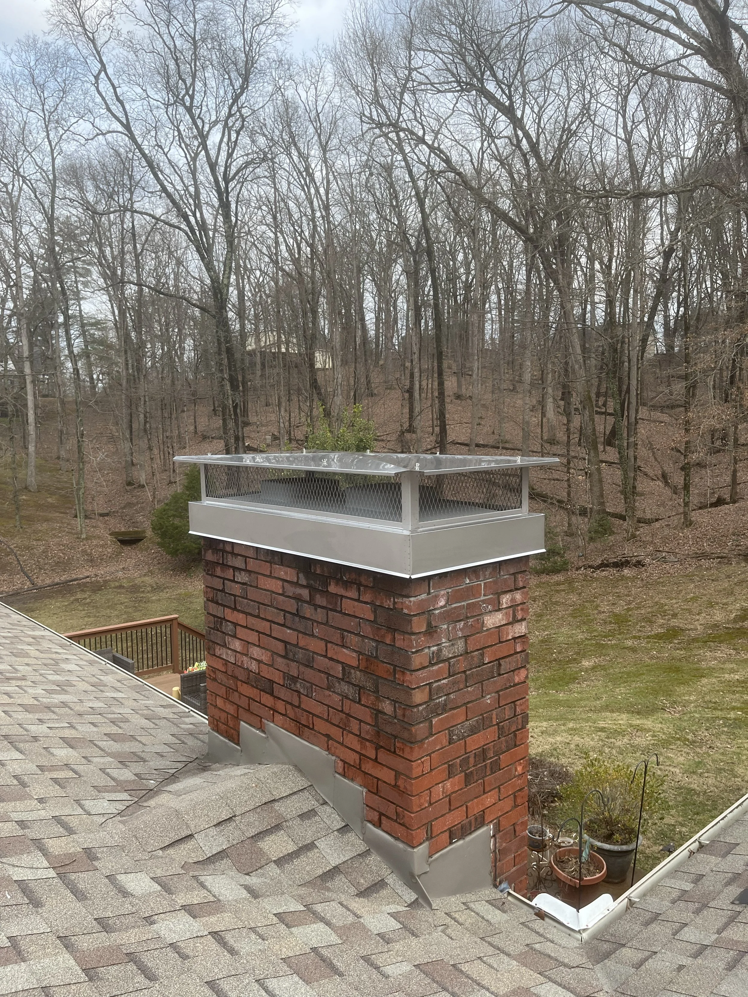 View of a brick chimney with a metal cap above it, attached to a brick wall on a house roof, with rooftops, trees without leaves, and a cloudy sky in the background.