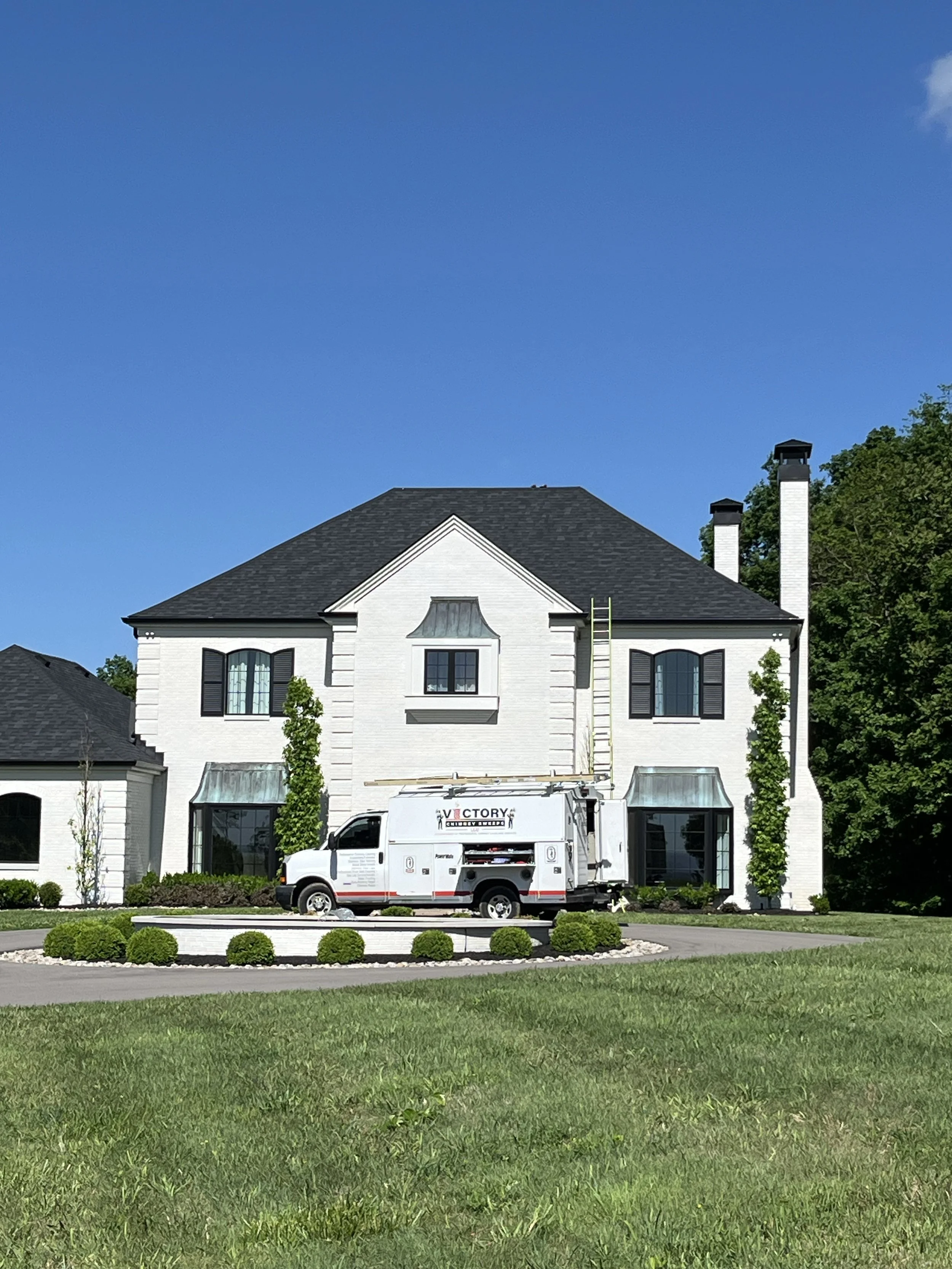 A white house with black shutters and a dark gray roof, two chimneys, and a front yard with green grass and small bushes. A white utility van parked in front of the house has the logo 'Victory' and construction equipment on its side.