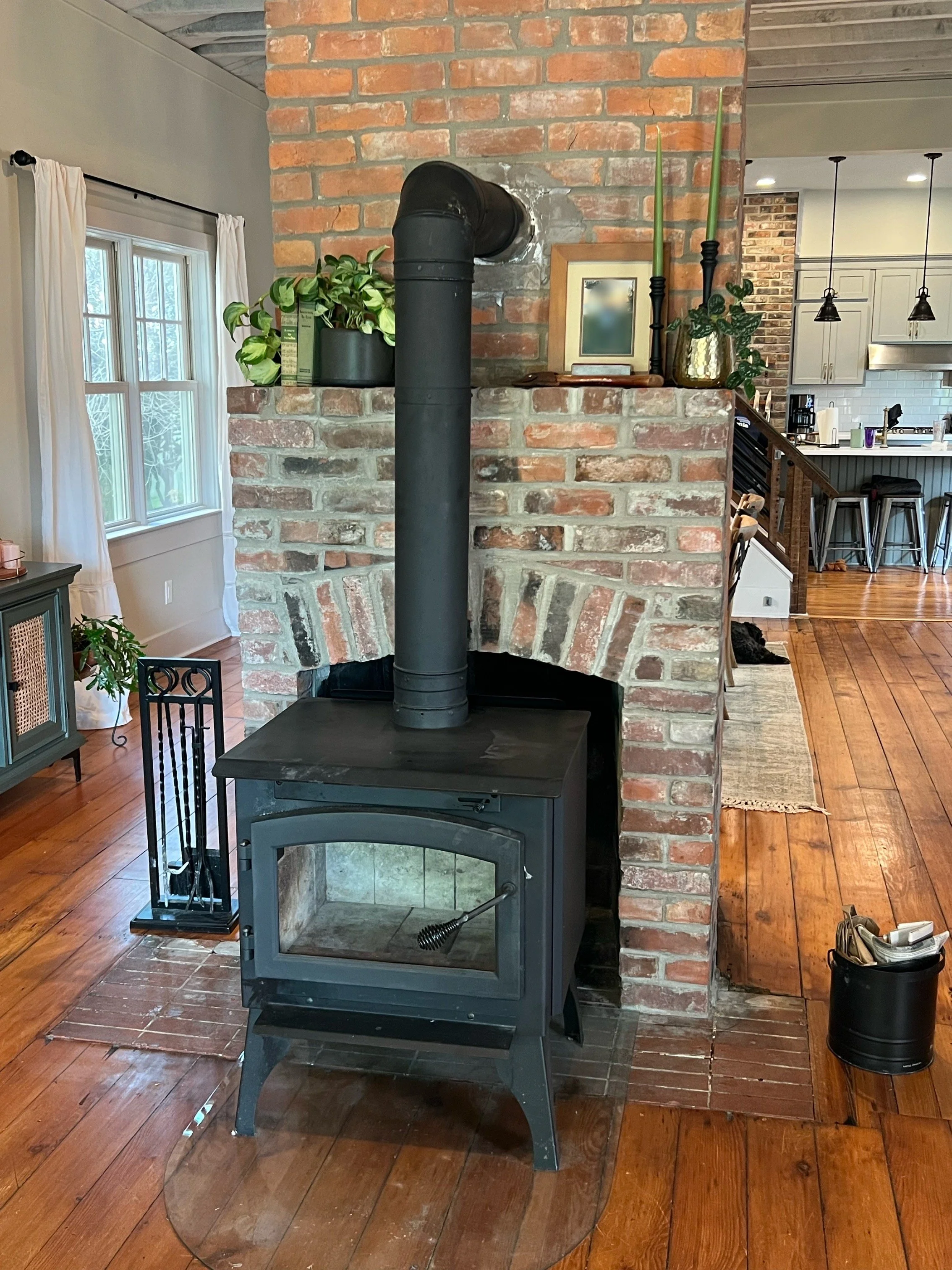 A black wood stove with a chimney pipe on a brick fireplace in a cozy living room. The room has hardwood floors, a window with white curtains, and various plants and furniture.