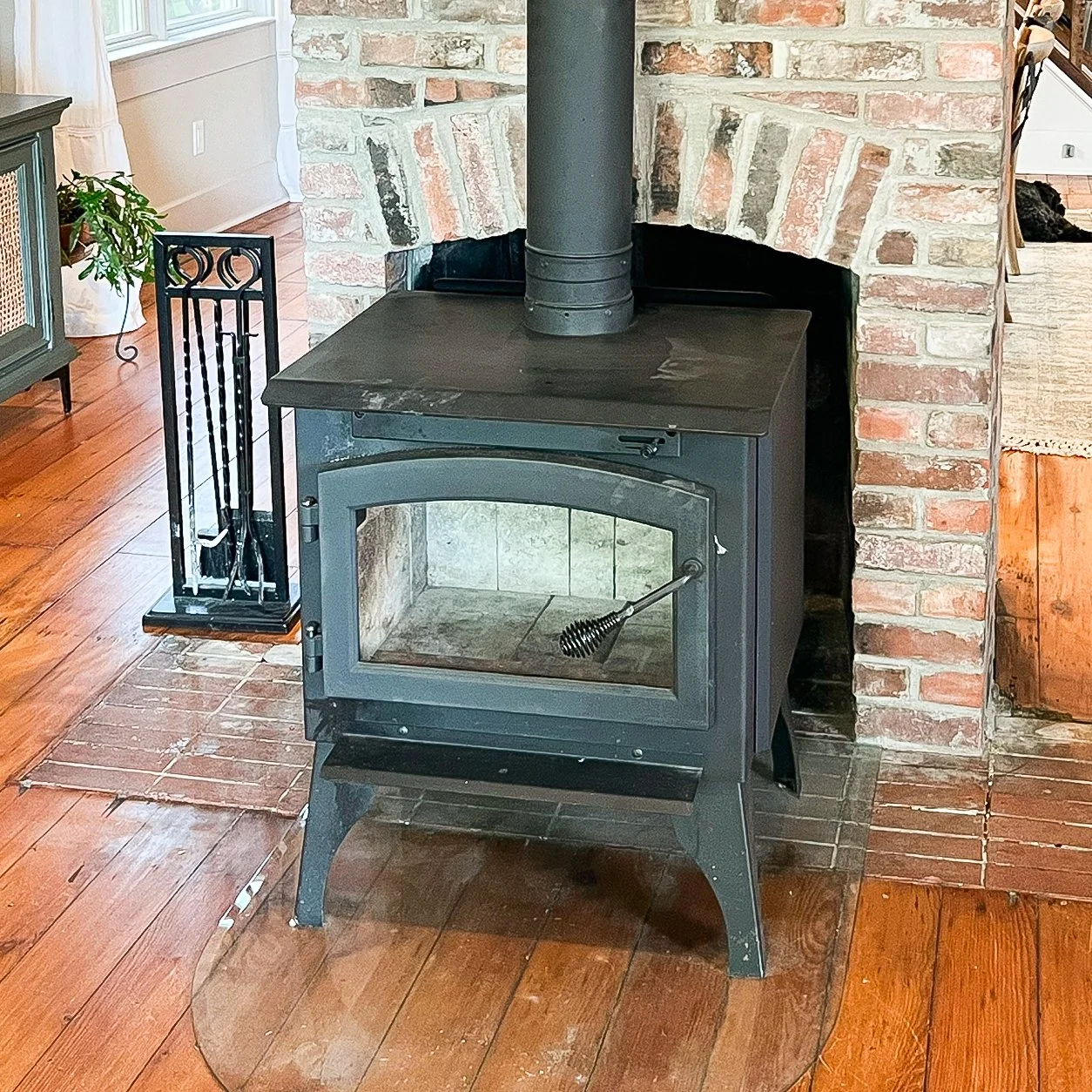 A black wood-burning stove with a glass door, placed on a brick hearth in front of a brick fireplace. There is a chimney pipe extending from the stove upward. To the left, a black metal tool holder and a handle with a brush are visible. Parts of wooden floors and a green cabinet are also visible in the background.