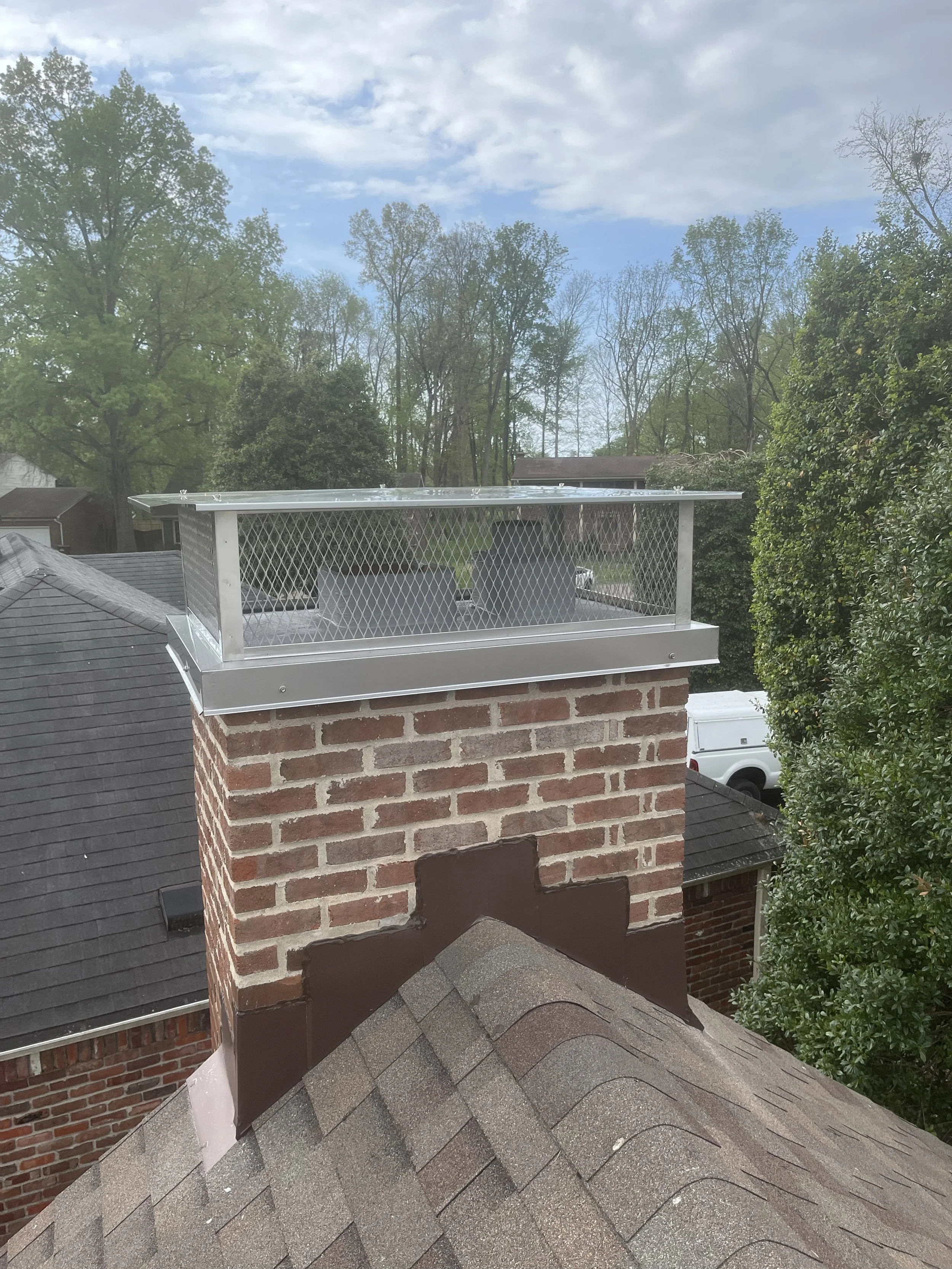 A brick chimney with a metal cap on top, situated on a sloped shingled roof, with trees and houses in the background under a partly cloudy sky.