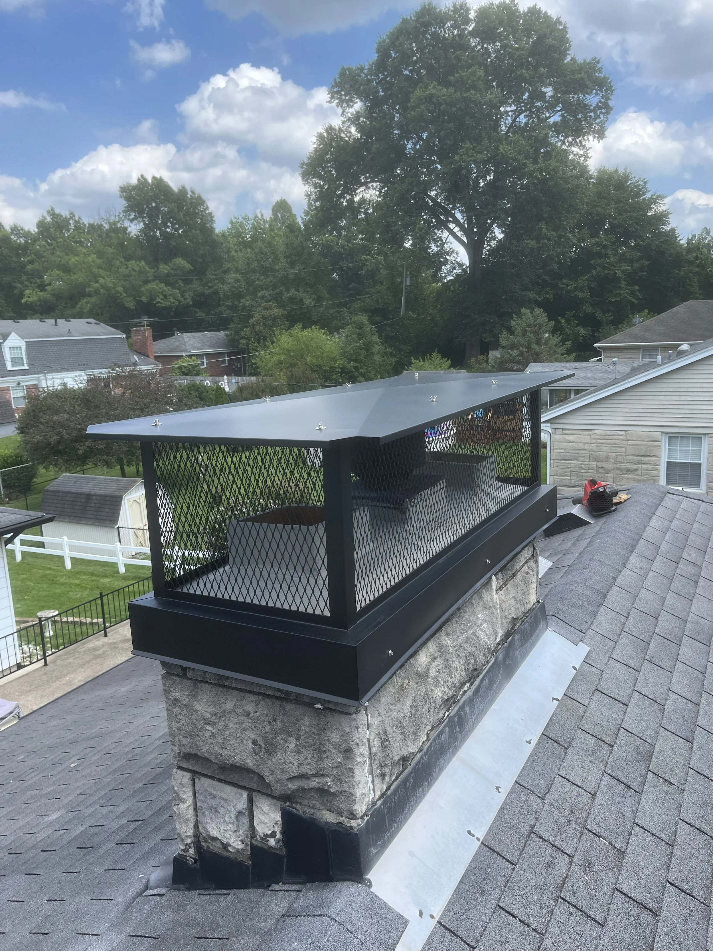View of a building's rooftop area with a chimney and a mountain of large stones supporting the structure. The stones are secured with a black metal cap and mesh railing. The rooftop has gray shingles, and residential houses and green trees are visibl