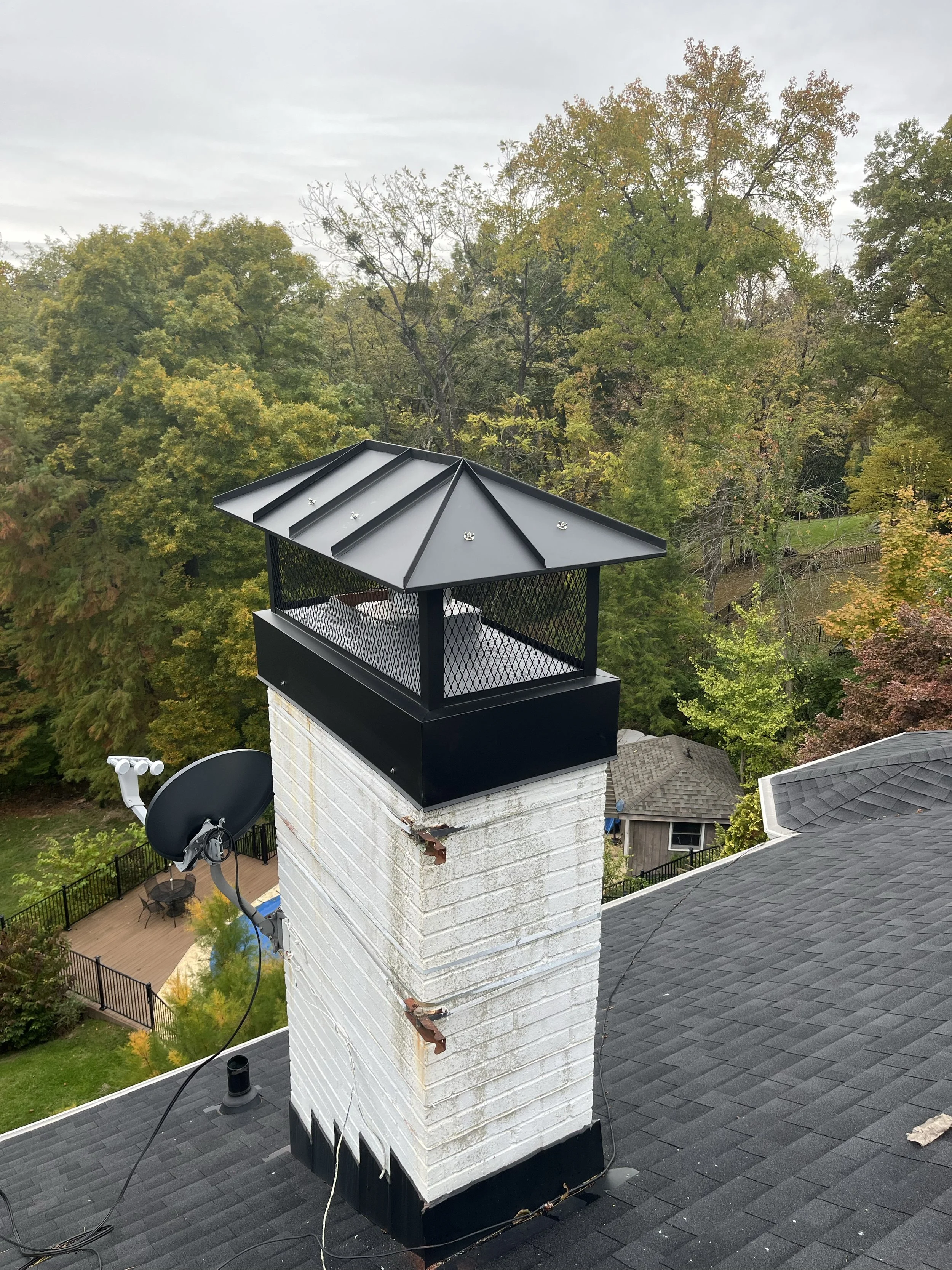 A chimney with a black metal cap on a house roof, with trees and cloudy sky in the background. There are satellite dish and cables visible near the chimney.