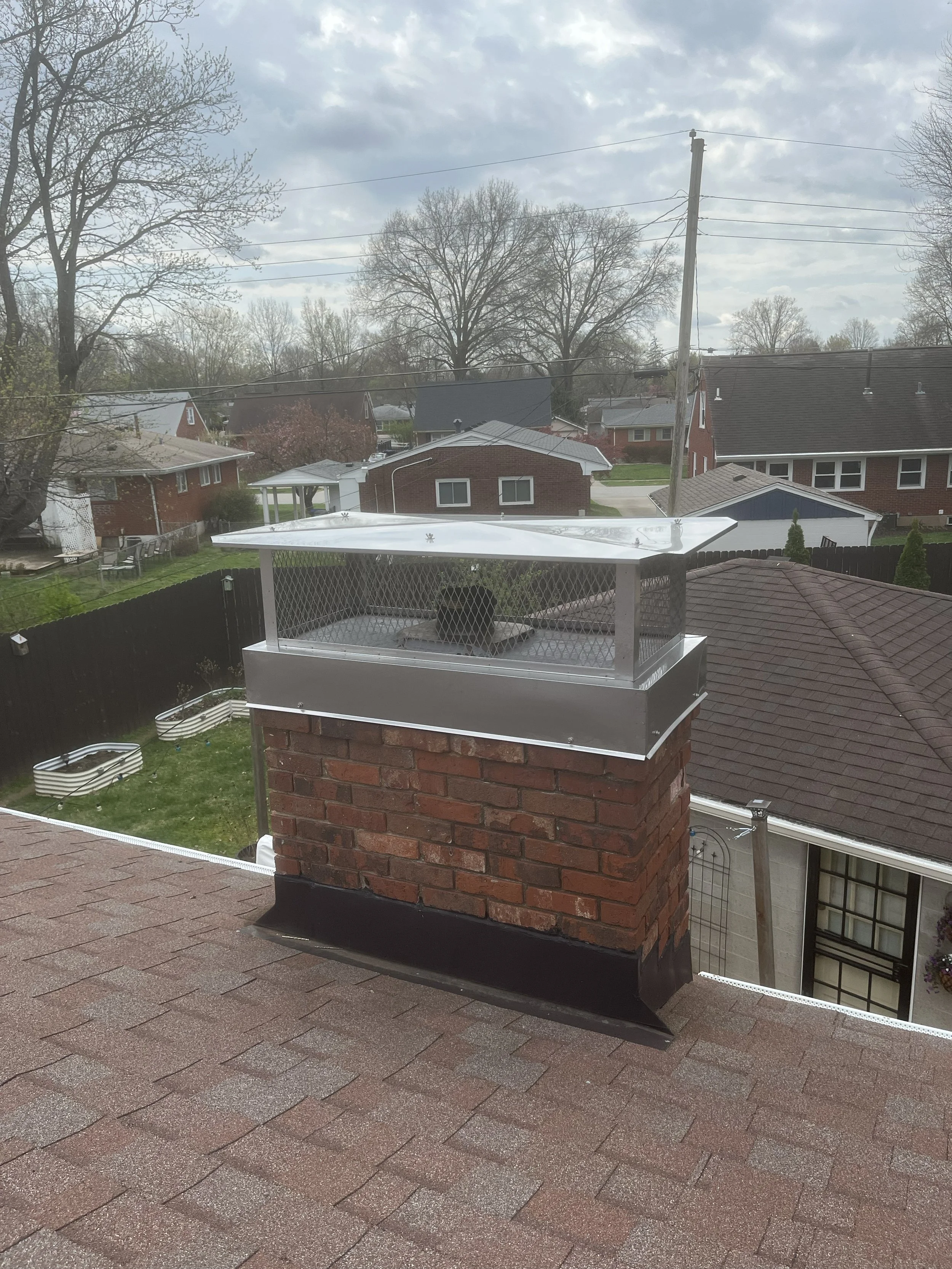 View of a residential neighborhood with houses, trees, and cloudy sky, seen from a second-story balcony with a brick chimney in the foreground.