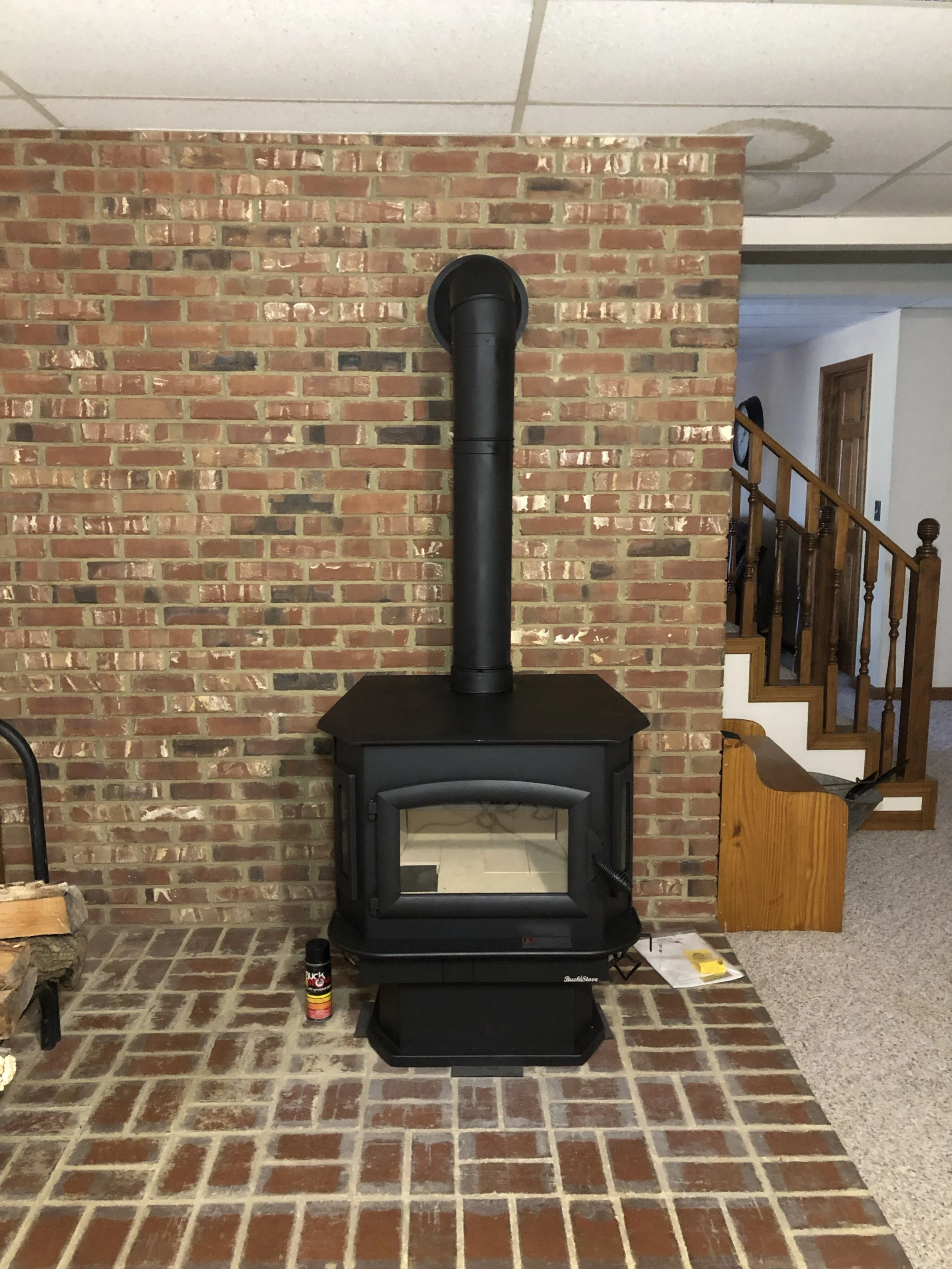 A living room with a brick fireplace, a black stove pipe, and various items on the floor, including a can of wood stain, some papers, and a yellow item, next to a wooden staircase with a brown handrail.
