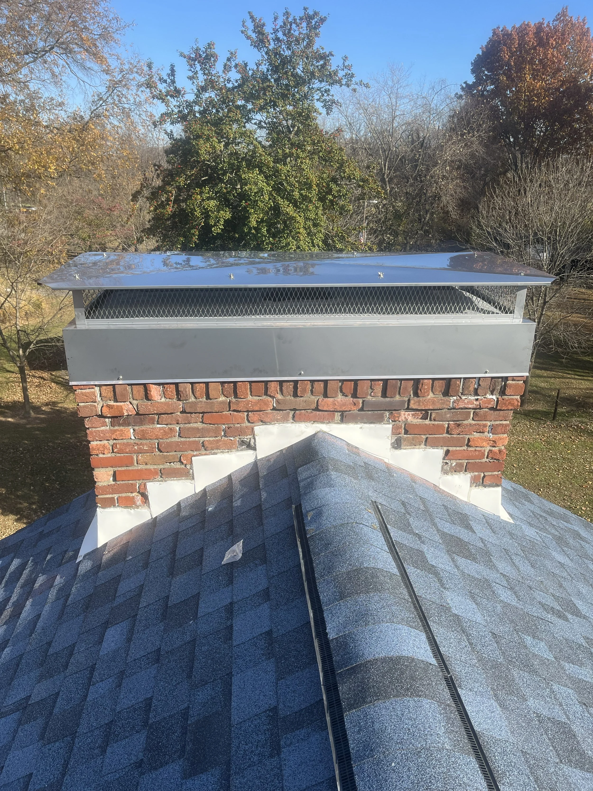 View of a brick chimney with metal cap and two black vent pipes on a residential roof, surrounded by trees with autumn foliage and a clear blue sky.