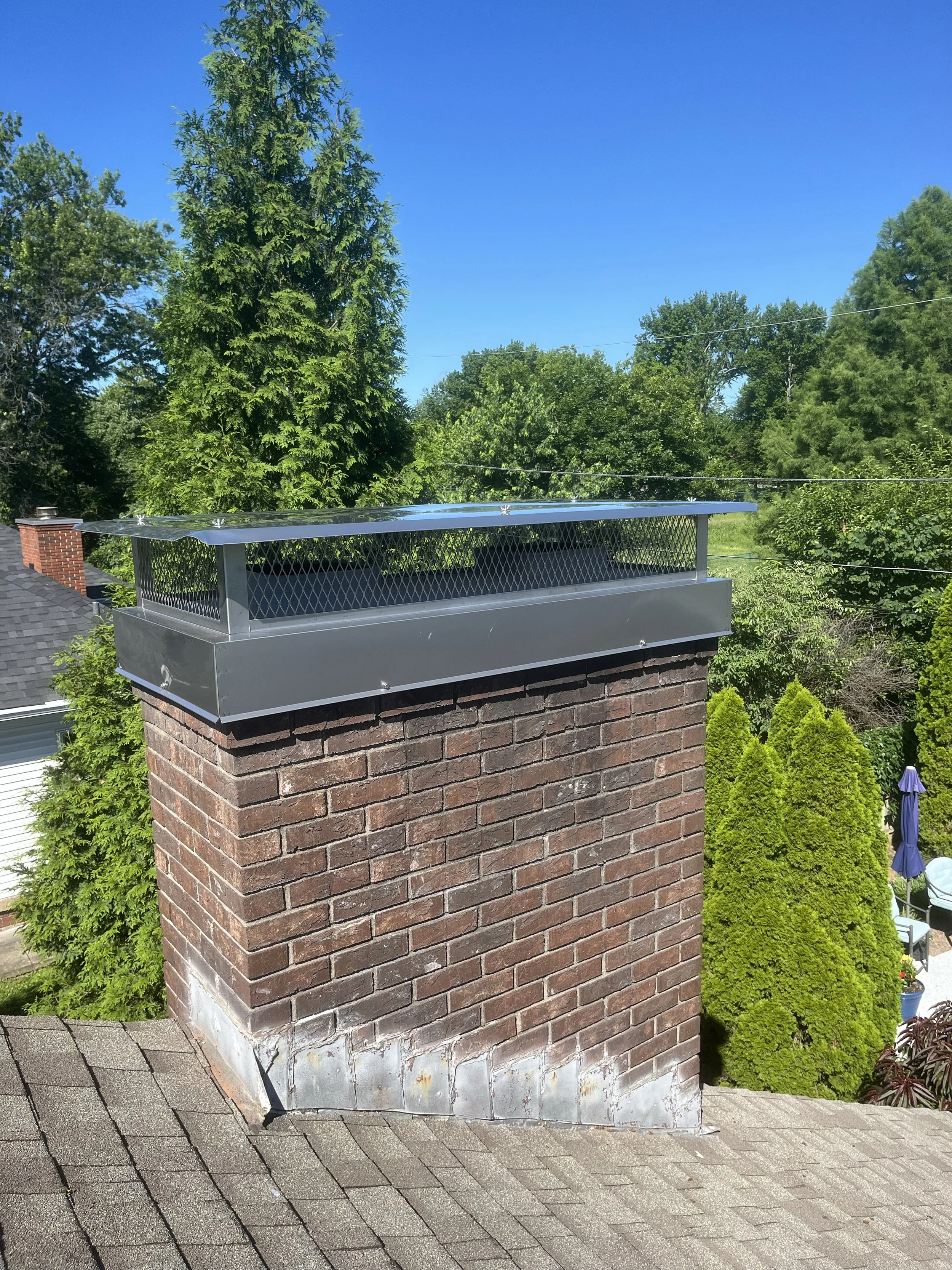 A brick chimney with a metal flashing roof cap on a sloped roof, surrounded by green trees and a clear blue sky.
