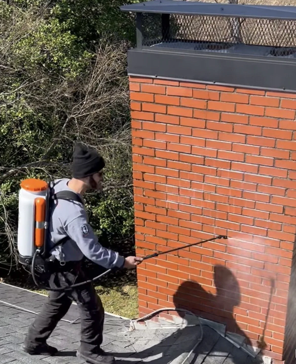 A man wearing a beanie and gray jacket using a sprayer to clean or treat a brick chimney outdoors.