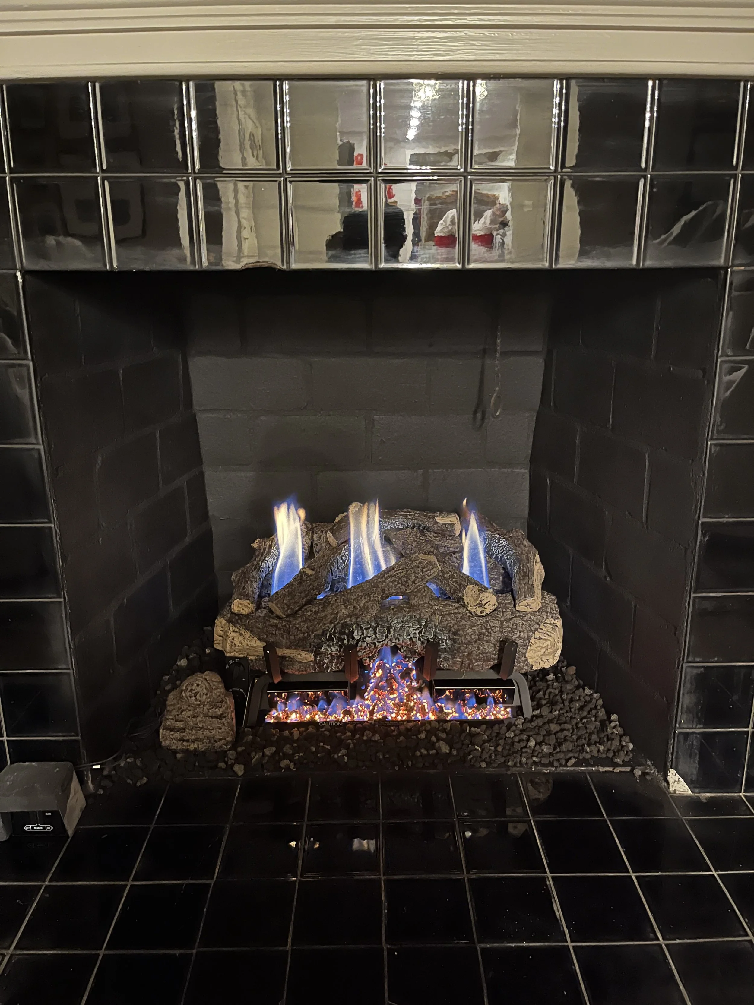 A modern gas fireplace with a black tile surround and a chrome mirrored mantel, showing blue and orange flames over rocks and wood logs inside.