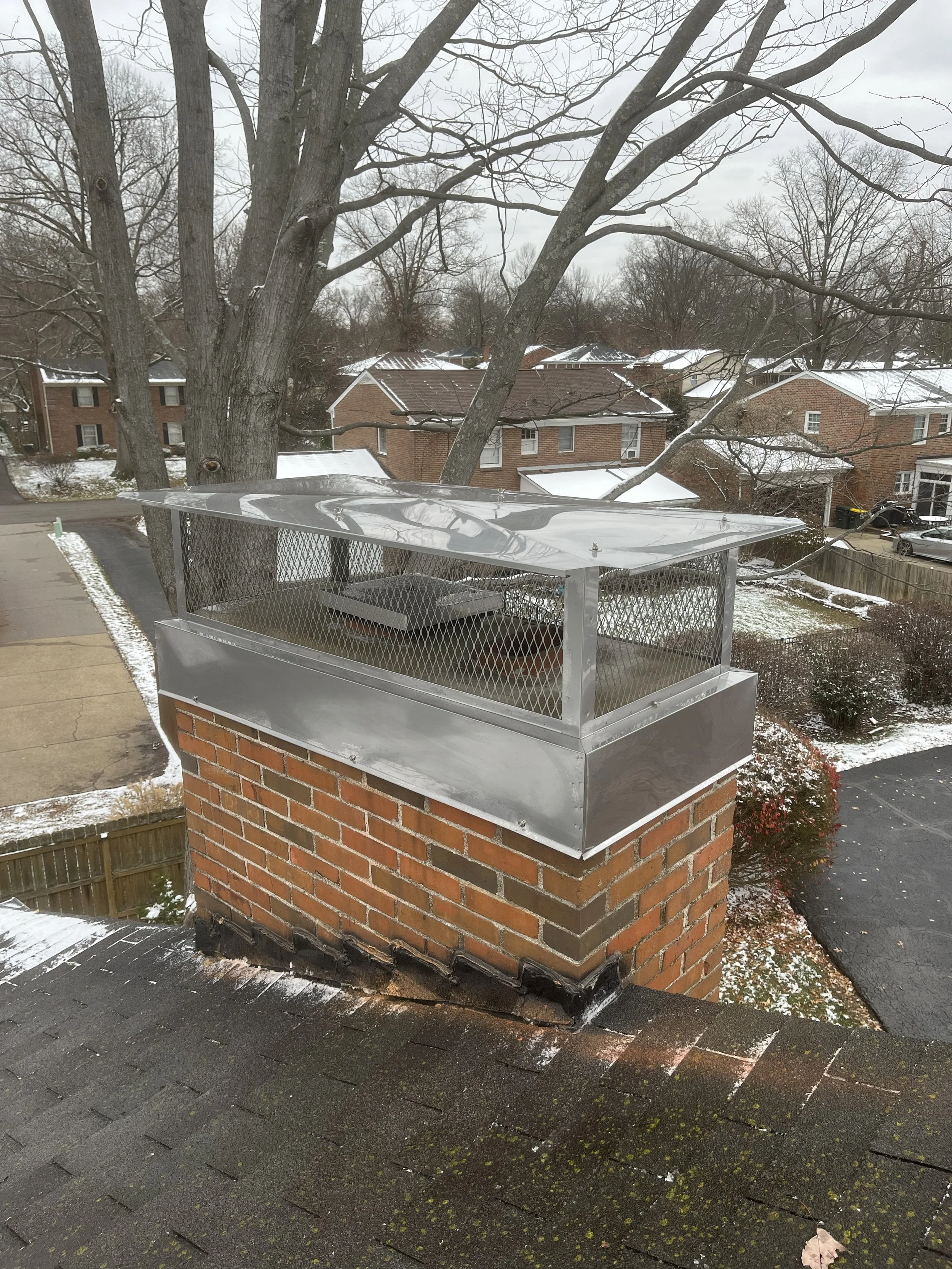 A rooftop with a brick chimney and a metal structure, possibly a vent or exhaust, on top of the chimney. There are snow patches on the roofs and ground, and bare tree branches in the background.