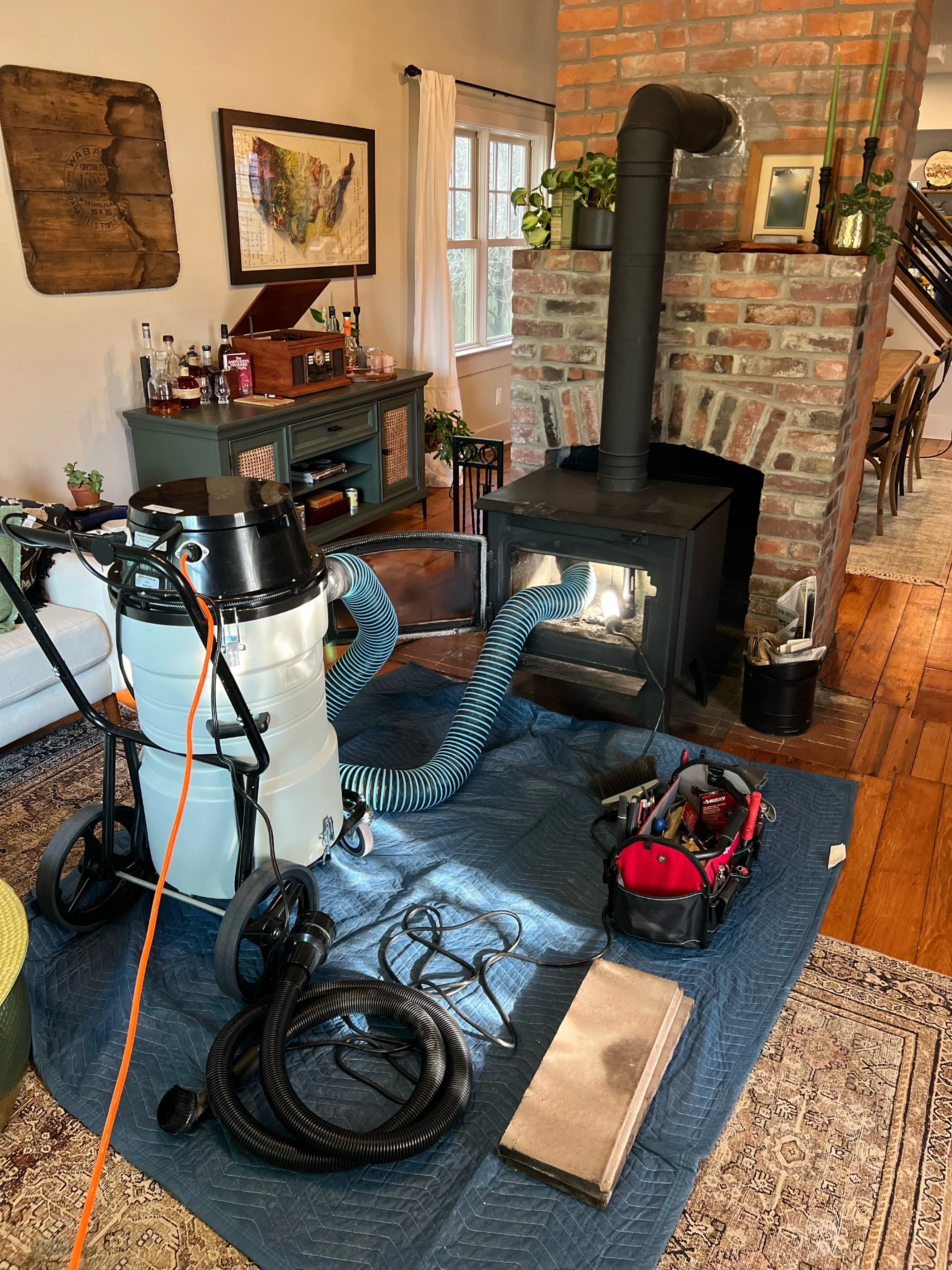 A room with a brick fireplace and a window, showing various tools and equipment on a tarp on the floor, including a vacuum, hoses, a red tool bag, and cleaning supplies.