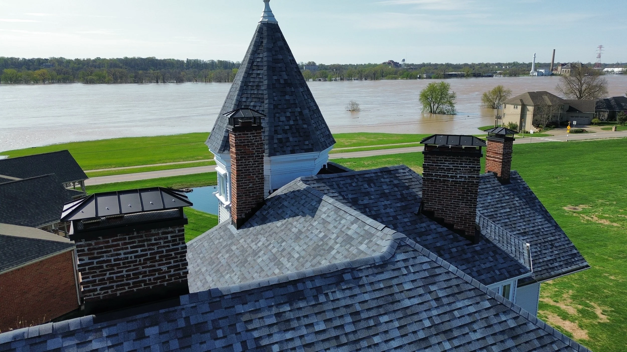 Roof shingles and chimneys of a house with a water flood in the background and trees partially submerged.