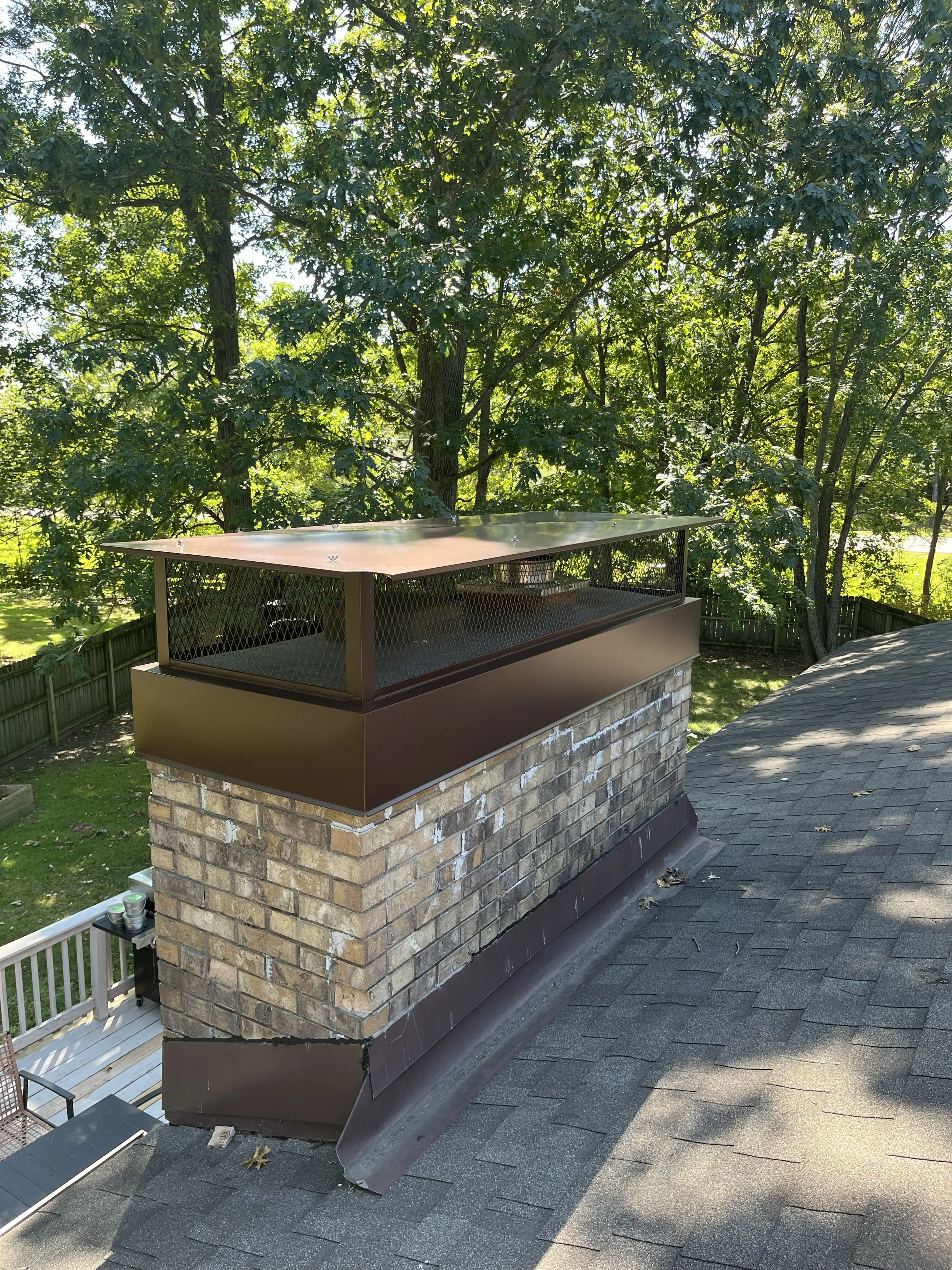 A house roof with a chimney and a flashing cap. The chimney is made of bricks with a brown metal cap. The roof has black asphalt shingles. There is a tree with green leaves in the background and a clear blue sky.