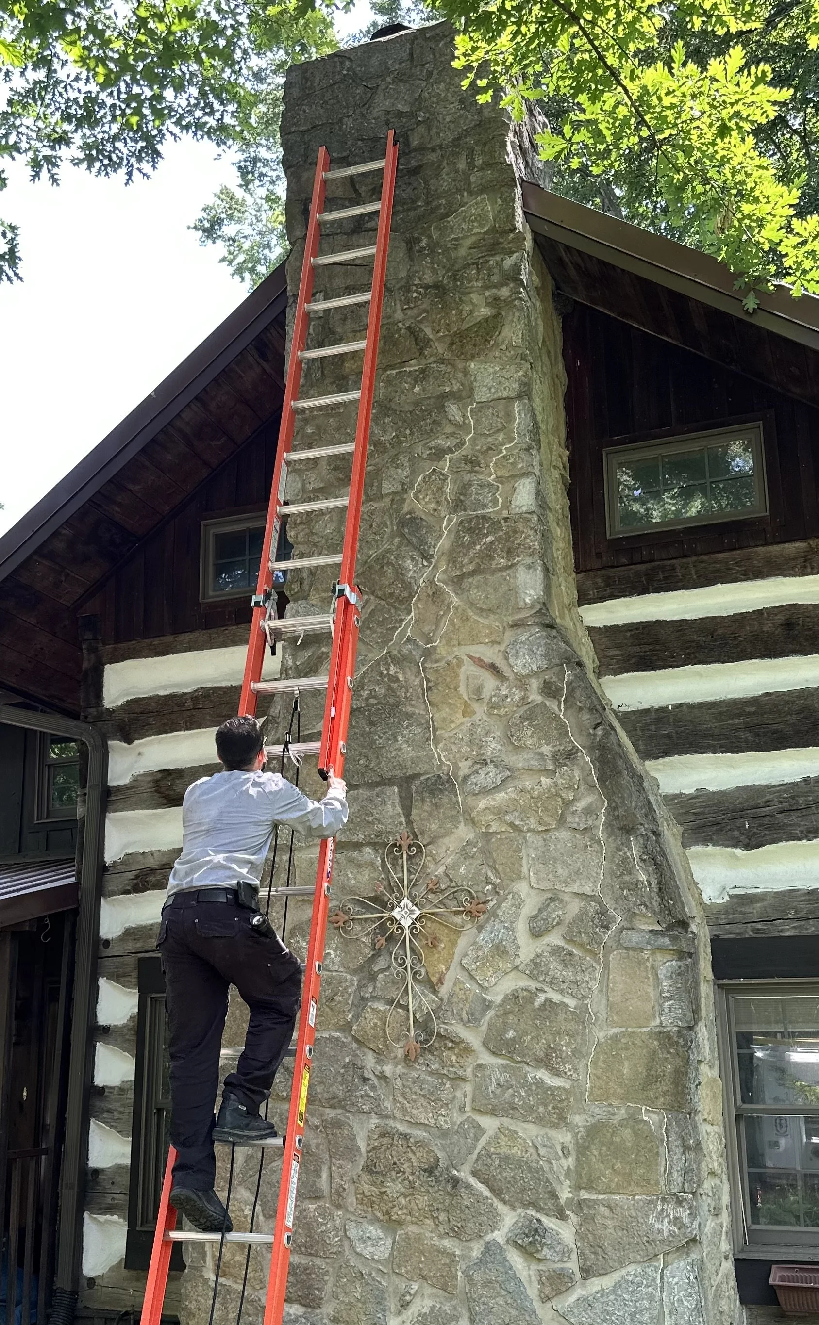 A person standing on a ladder fixing or inspecting a stone chimney on a house with a log cabin style exterior. The house has a black metal roof and a window, with greenery overhead.