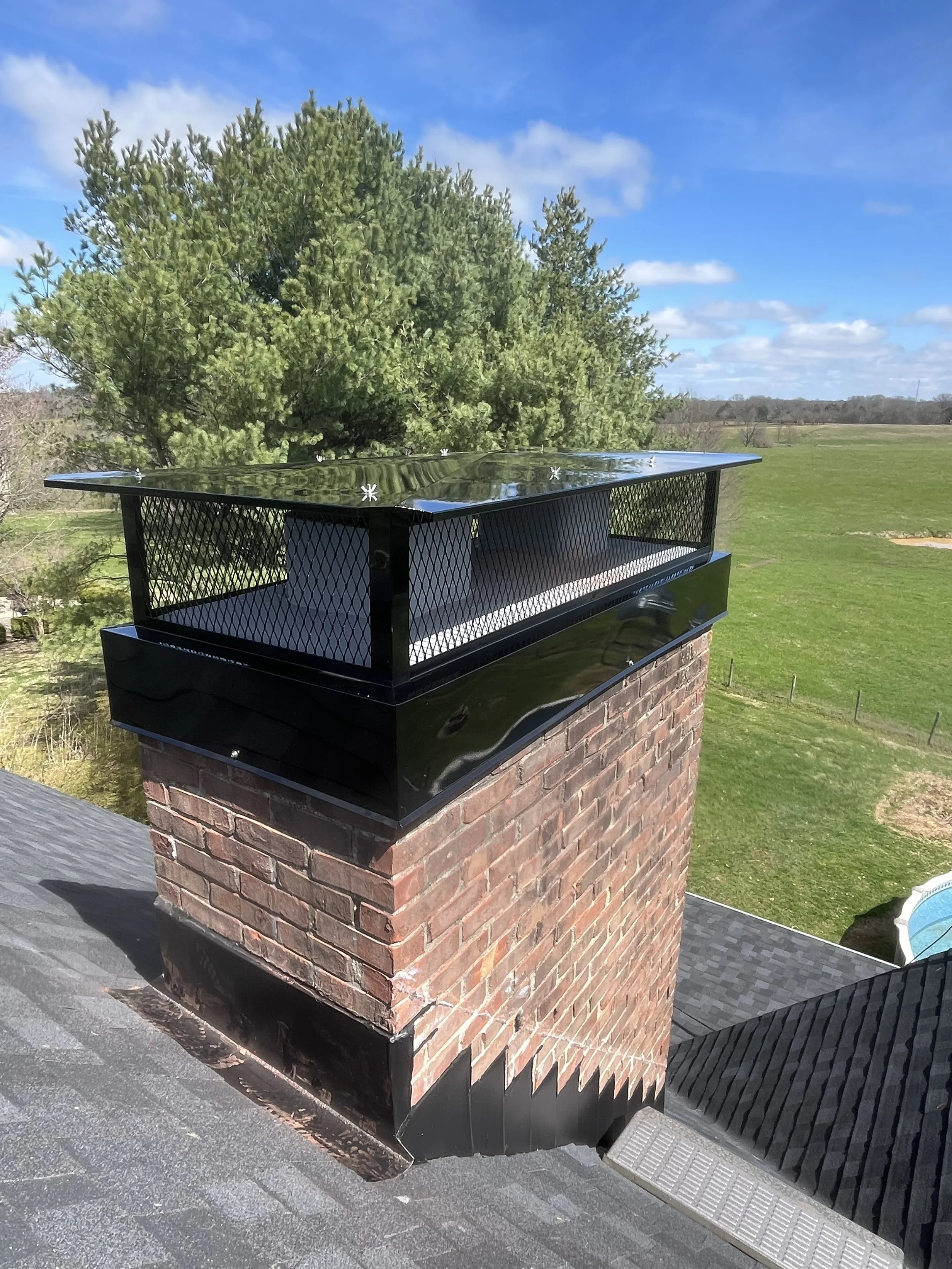 Photo of a brick chimney with a metal cap on top, located on a rooftop amid a grassy landscape with green trees and a partly cloudy sky.