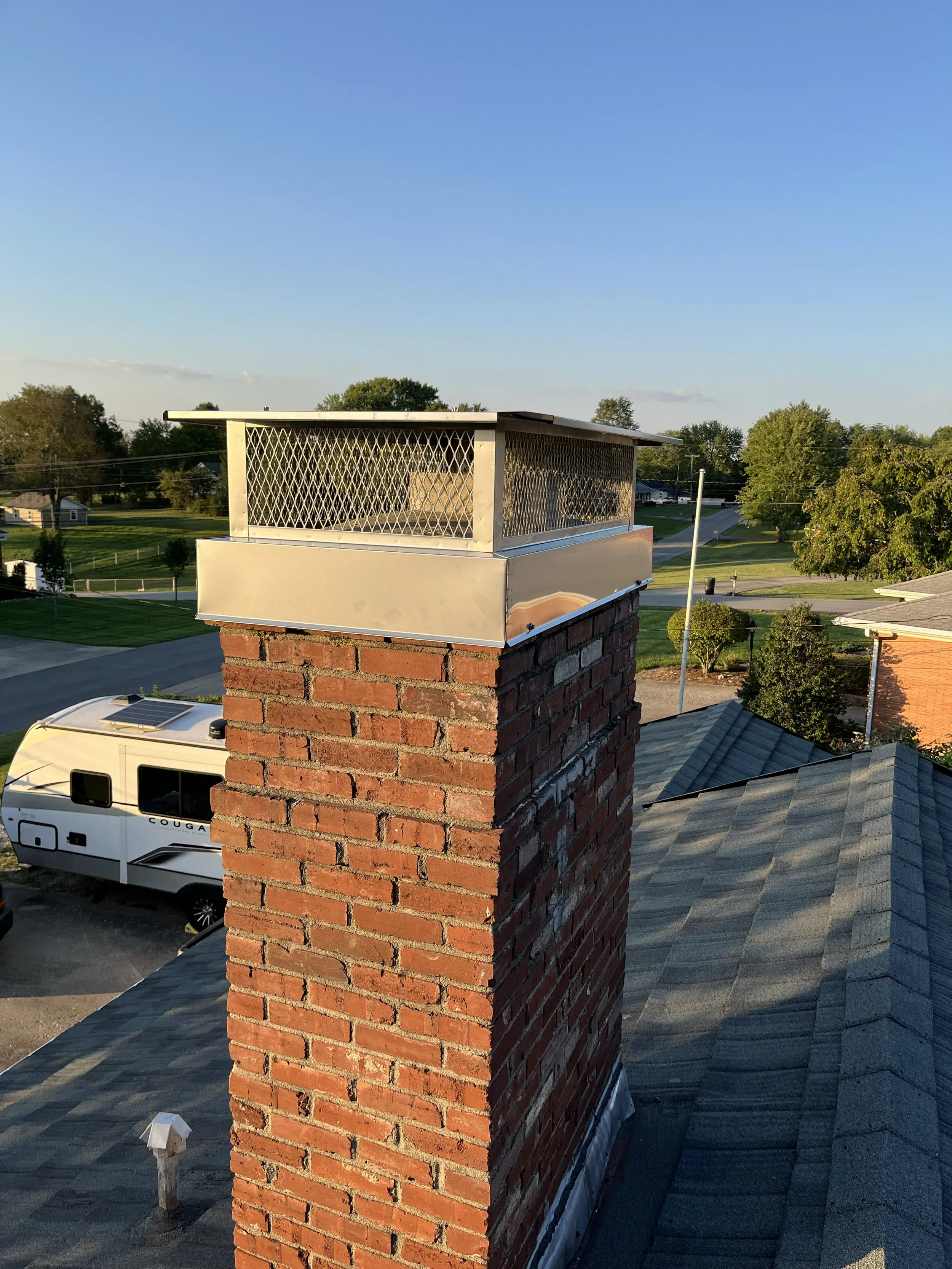 View from a rooftop showing a brick chimney with a metal cap, a vent pipe, nearby rooftops, trees, open sky, and a recreational vehicle parked below.