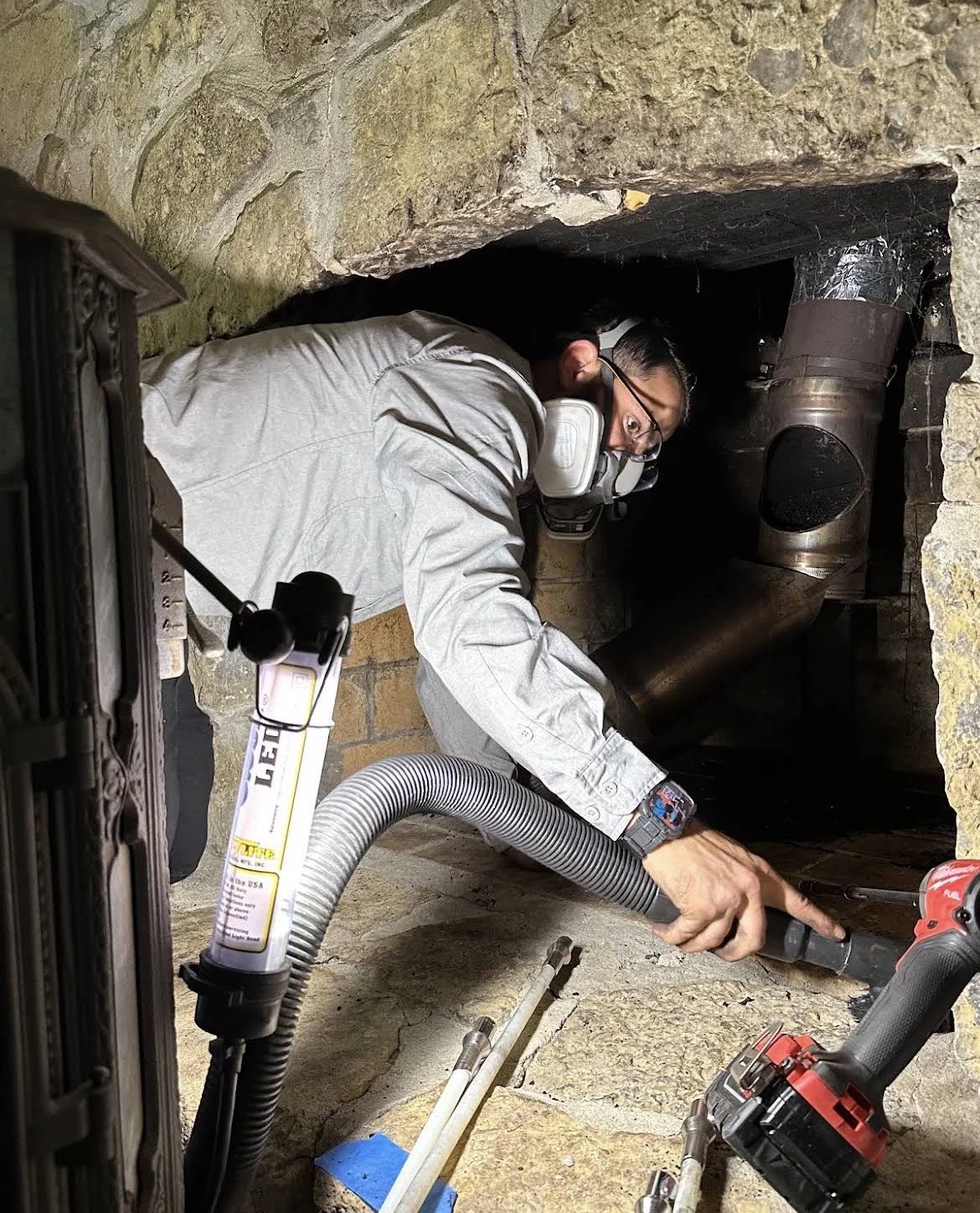 A man wearing a respirator mask and safety gloves working on plumbing inside a small, dark stone-lined crawl space or basement with pipes and tools around him.