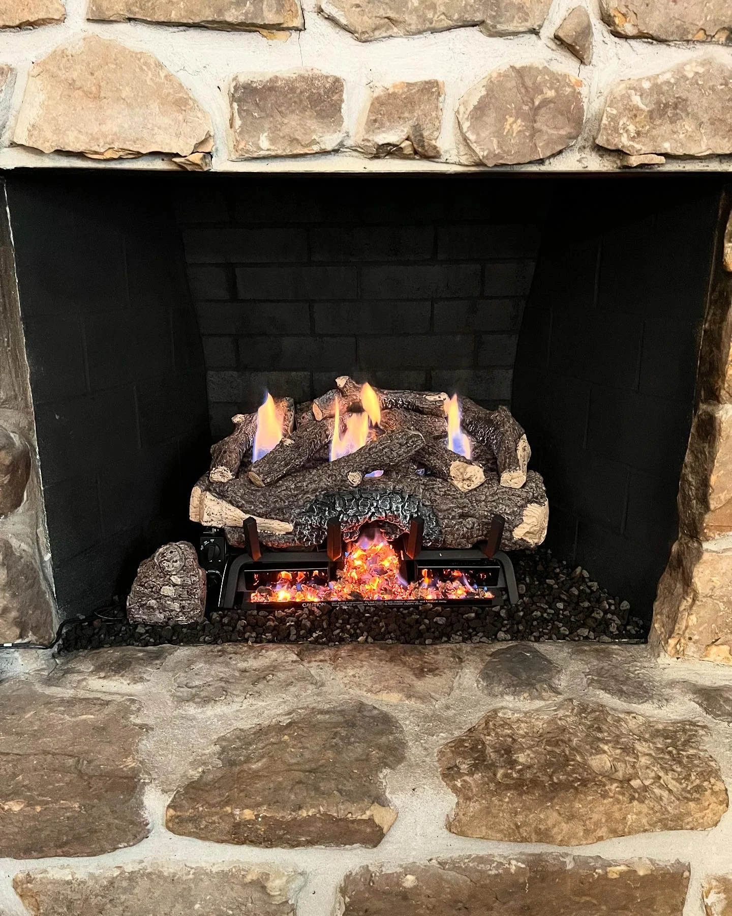 A cozy stone fireplace with a gas log fire, featuring faux logs and flames, surrounded by large beige and brown stones.