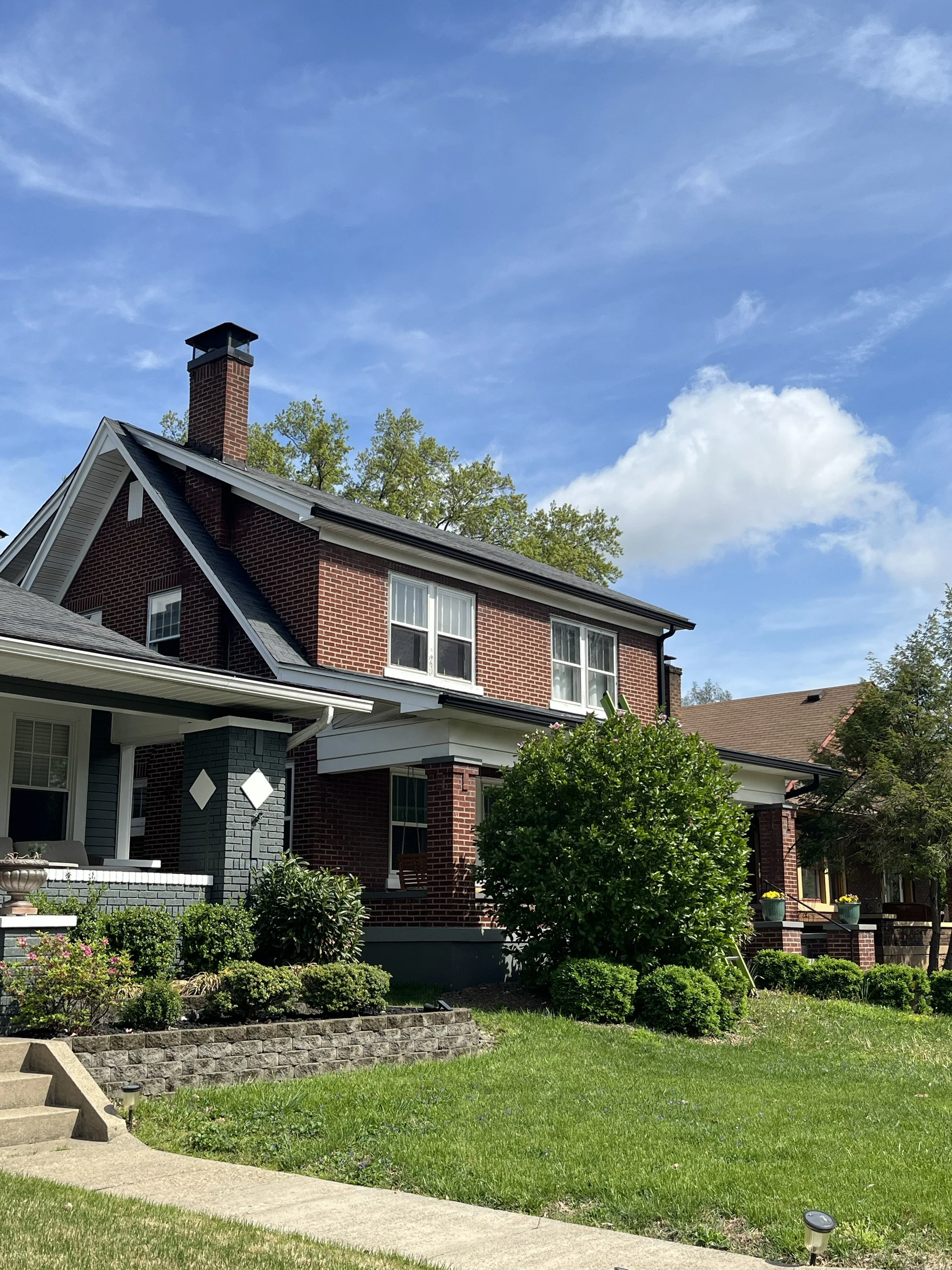 Front yard of a brick house with a landscaped garden, green lawn, pathway, and outdoor lighting under a partly cloudy blue sky.