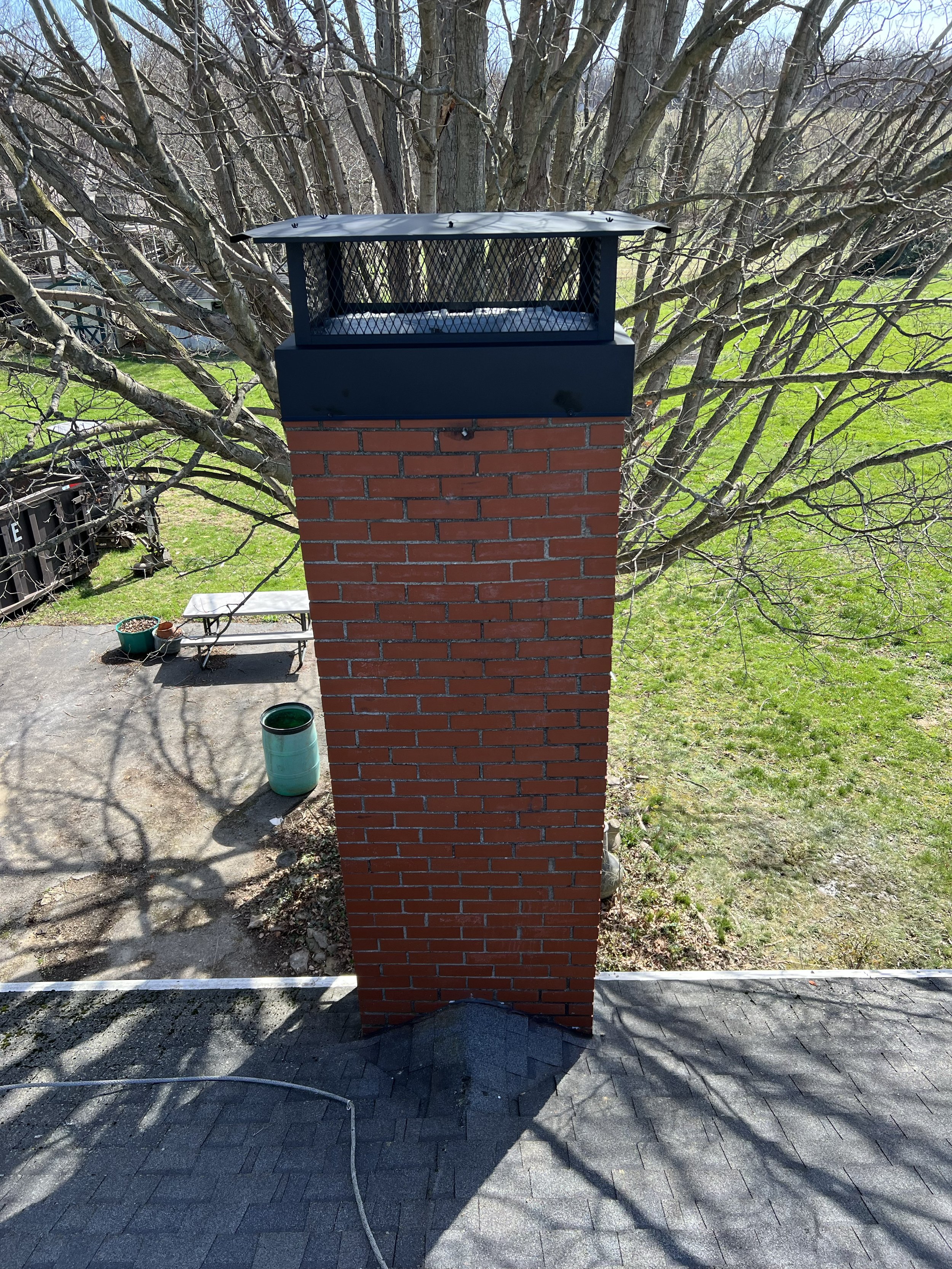 A brick chimney with a black metal cap on top, situated on a paved area outside, with a large, leafless tree in the background and a green lawn to the right.