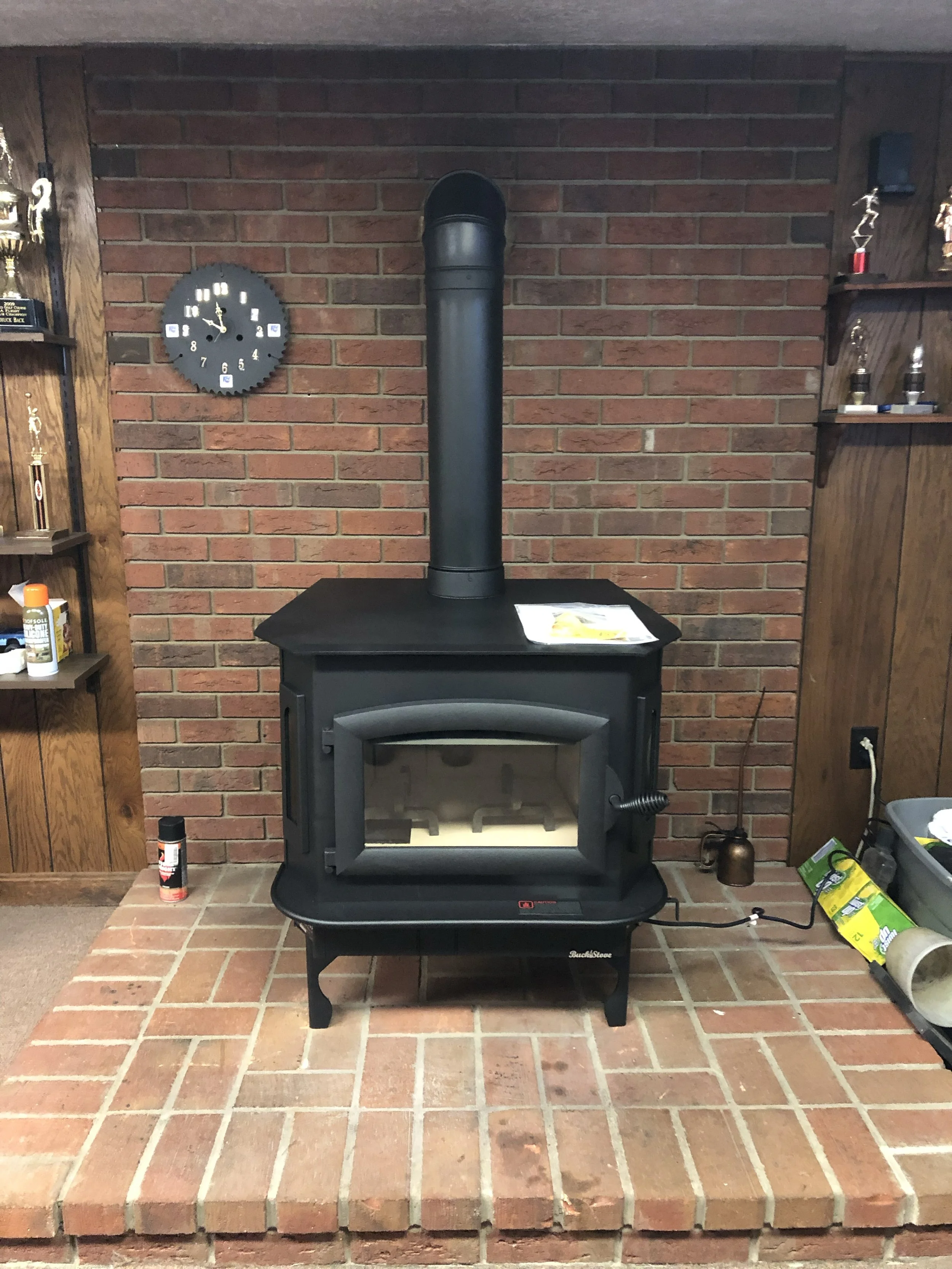 Black wood stove with a metal stovepipe in front of a brick wall, with trophies and shelves on either side, and various items on the floor.