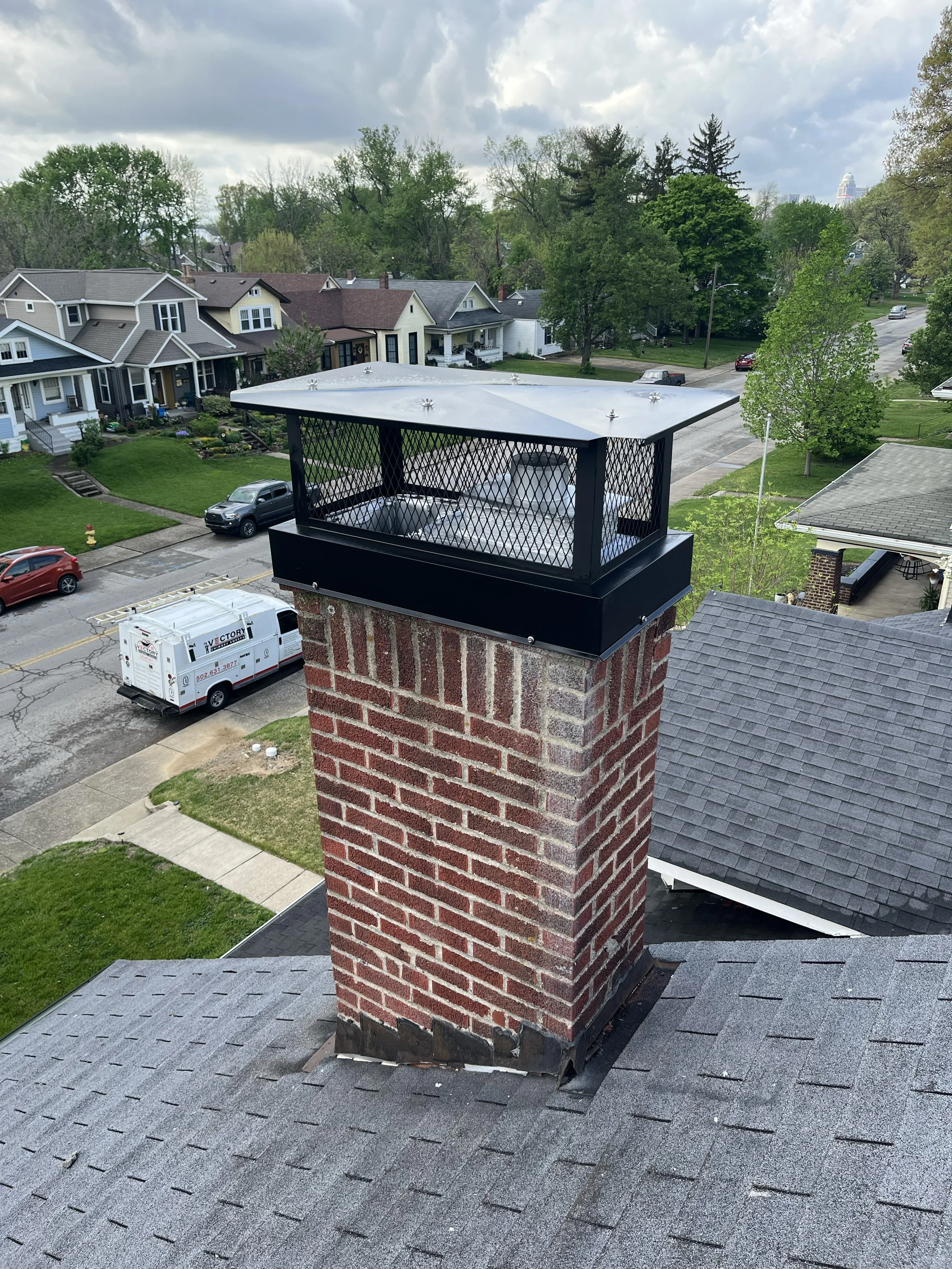A rooftop chimney with a cap and a metal shield on a shingled roof of a house, with a residential neighborhood, trees, and cloudy sky in the background.