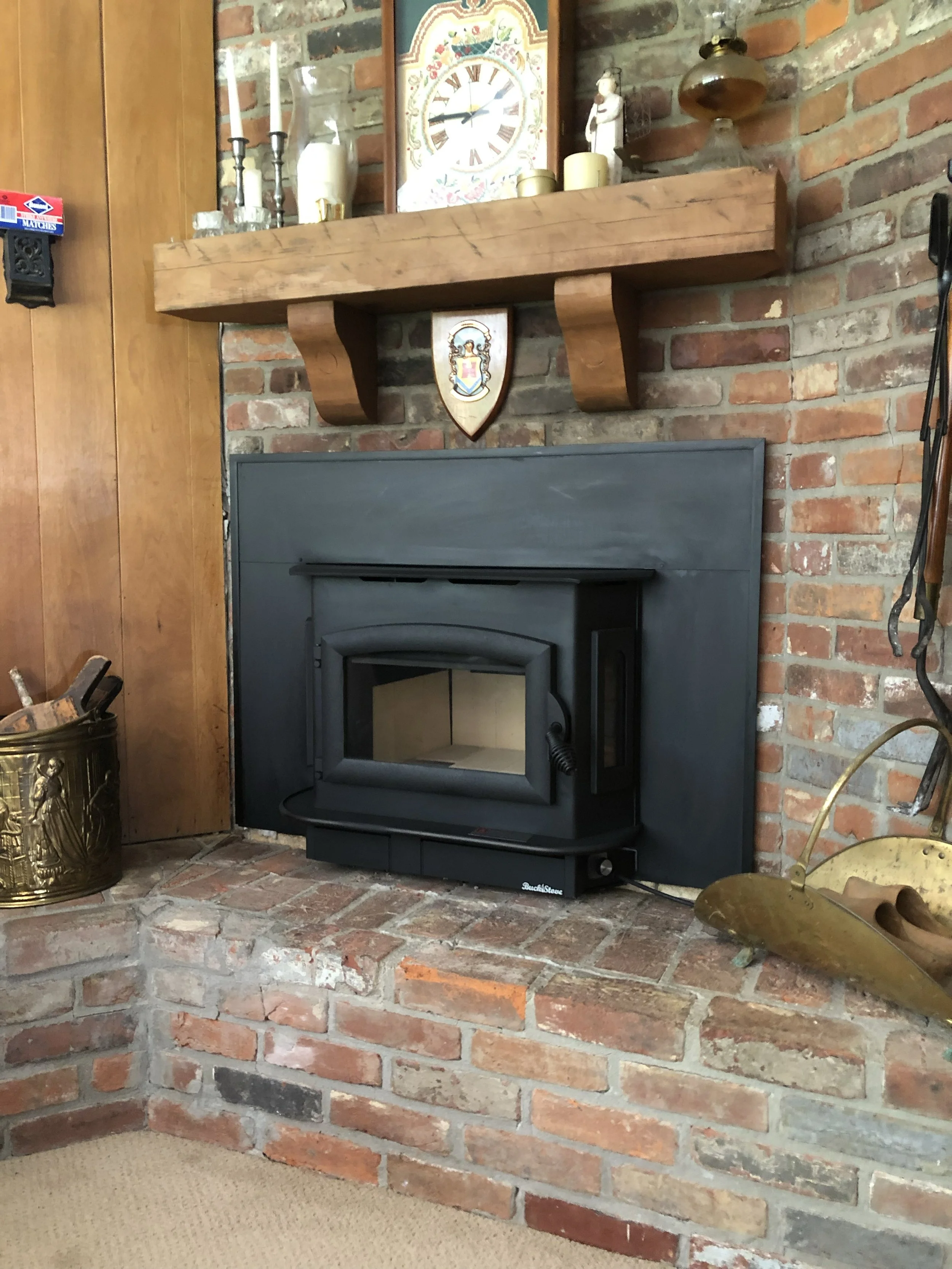 A cozy interior fireplace scene featuring a black wood stove set into a brick fireplace. Above the stove is a wooden mantel decorated with various items including candles, a clock, vases, and small figurines. The brick wall behind the mantel adds war
