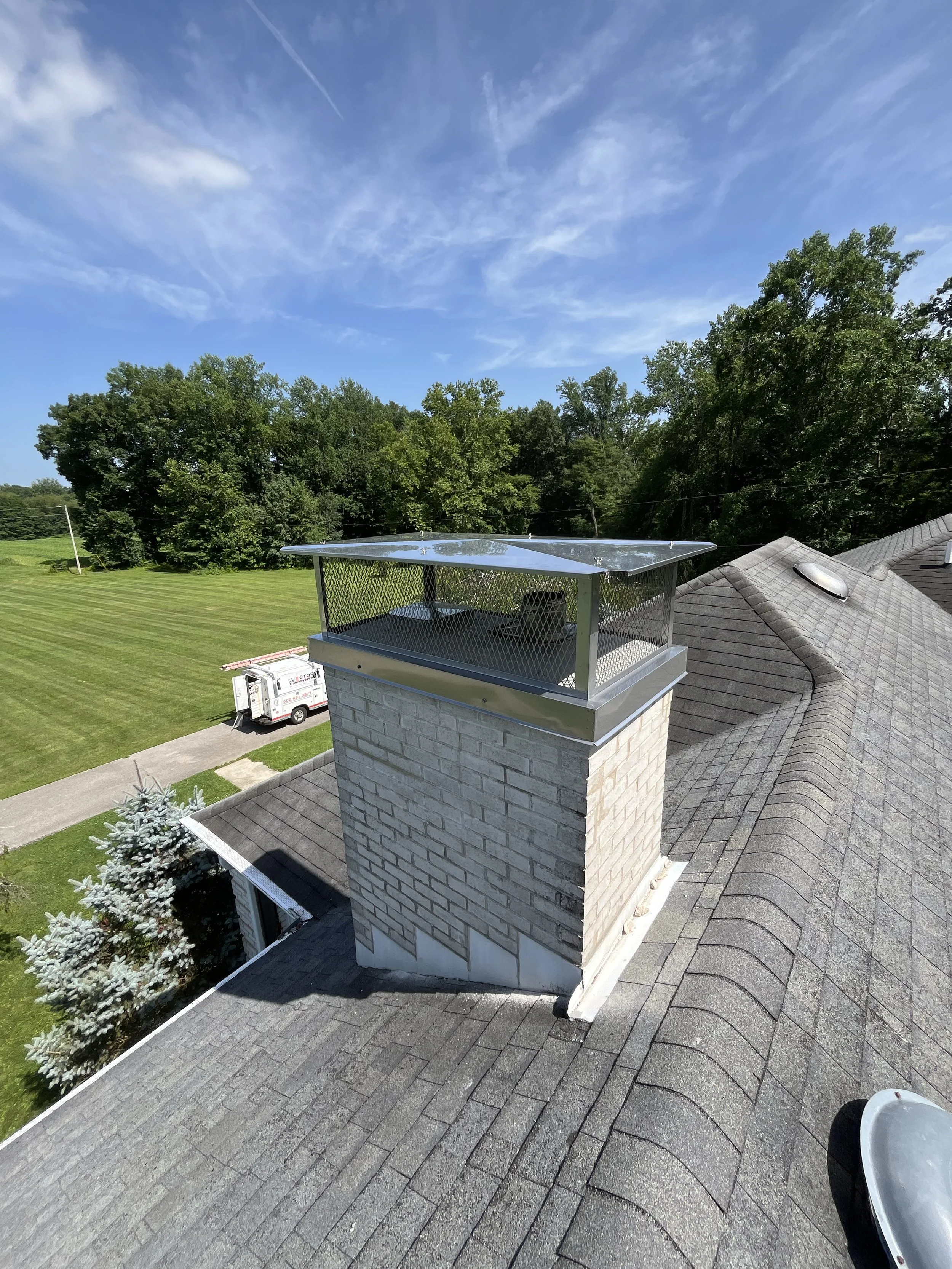 View of a rooftop with a brick chimney and metal chimney cap, shingles, a grill, and a backyard with green grass and trees under a blue sky with clouds.