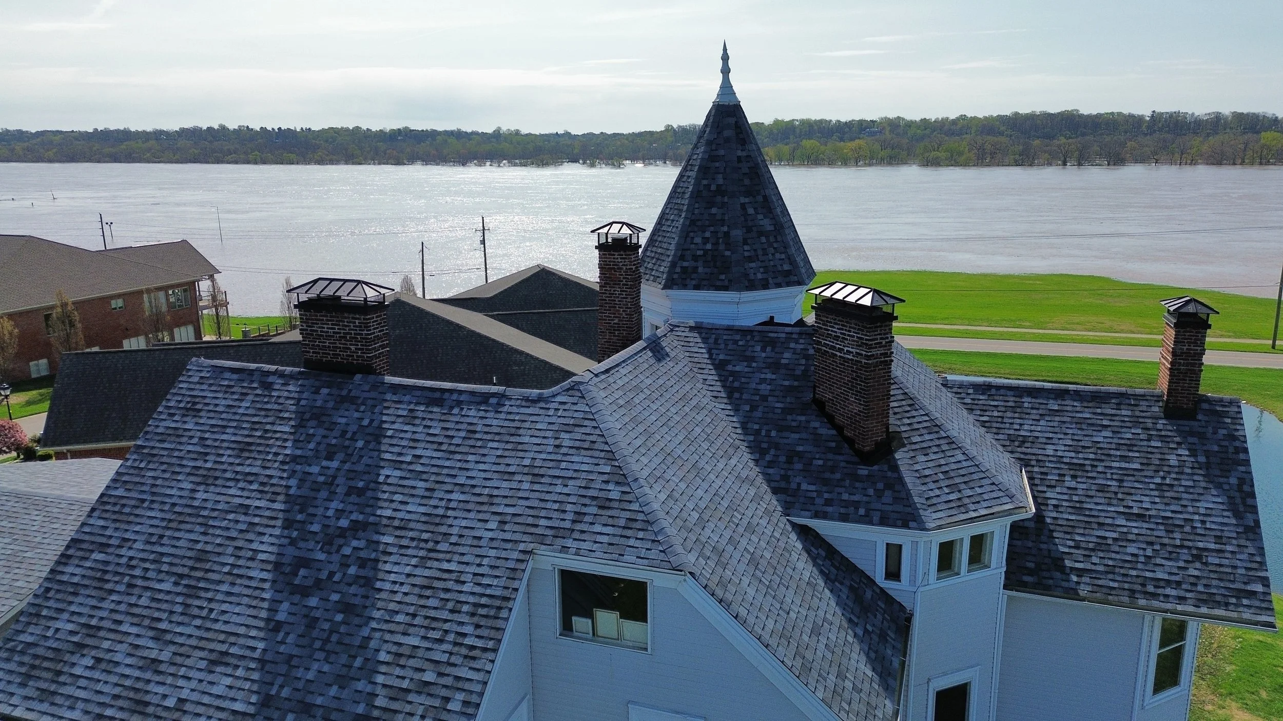 Aerial view of a house with multiple rooftops and a prominent turret, situated near a large body of water with a distant treeline.