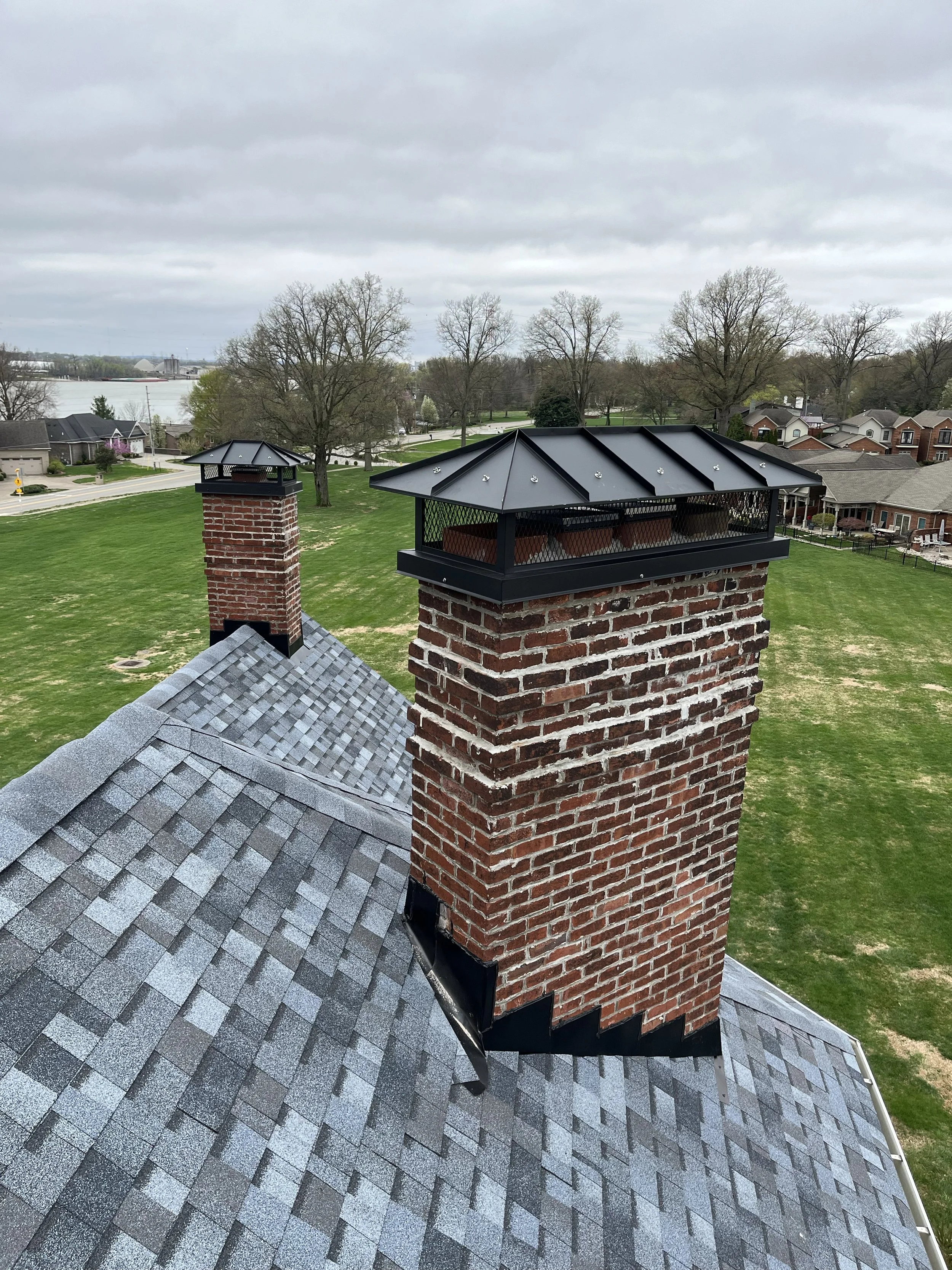 View of a house roof with a chimney topped by a black metal cap, and a metal chimney flashing around the brick chimney, with a background of trees, part of a city, and a river under a cloudy sky.