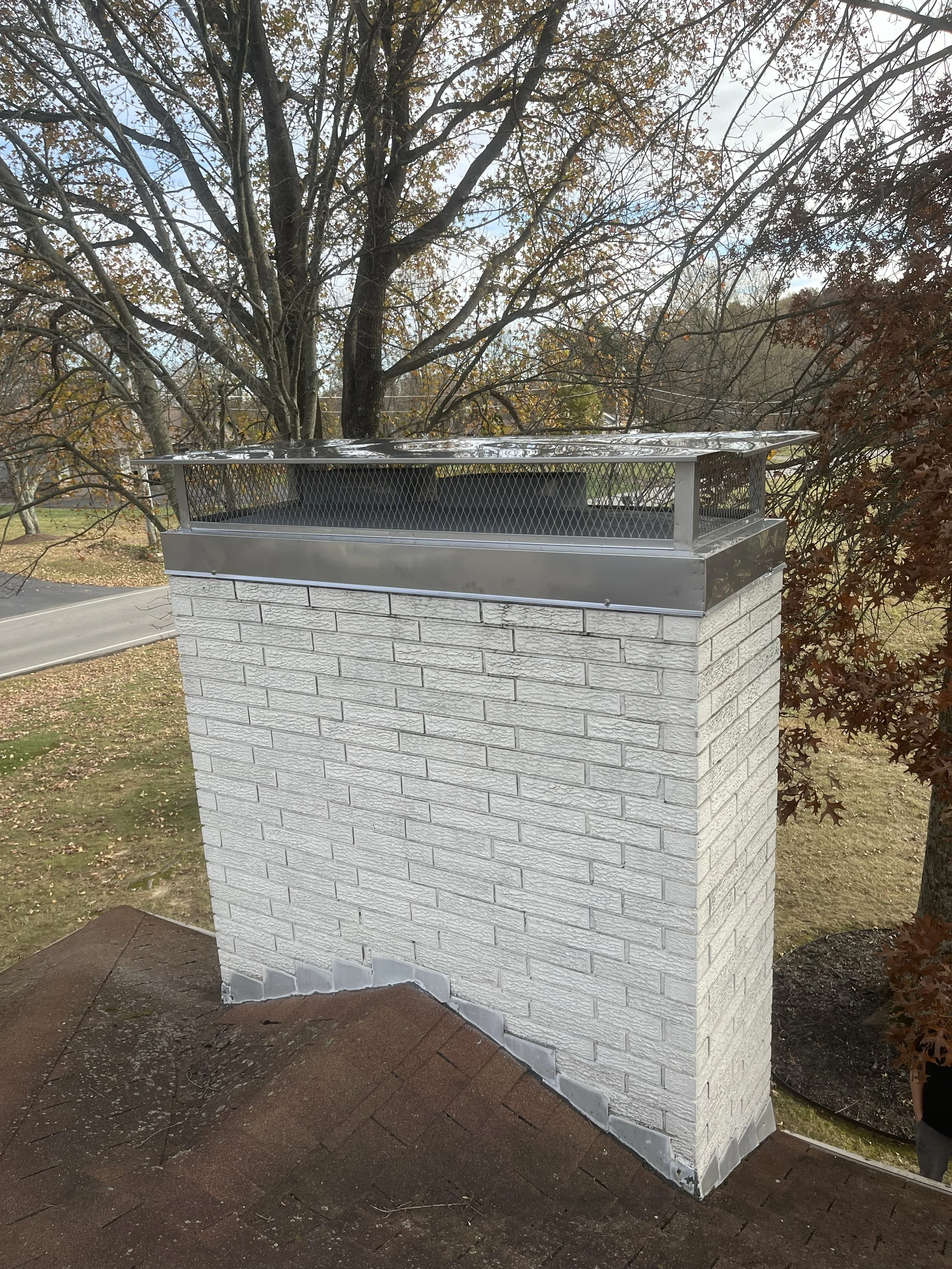 A brick chimney with a metal cap and a wire mesh enclosure on top, situated on a roof with brown shingles. In the background, trees with autumn leaves and a cloudy sky are visible.
