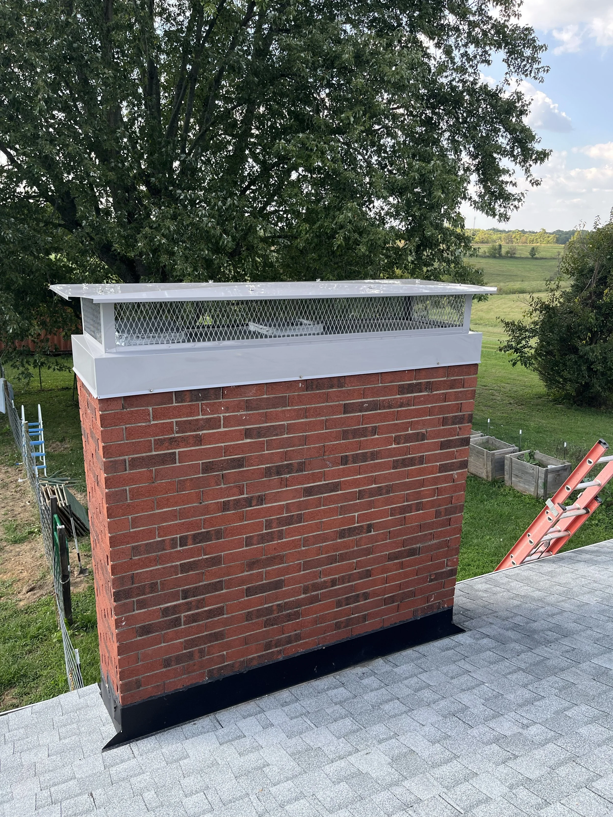 Brick chimney with metal cap and mesh vent on roof, overlooking green fields and trees under a partly cloudy sky.