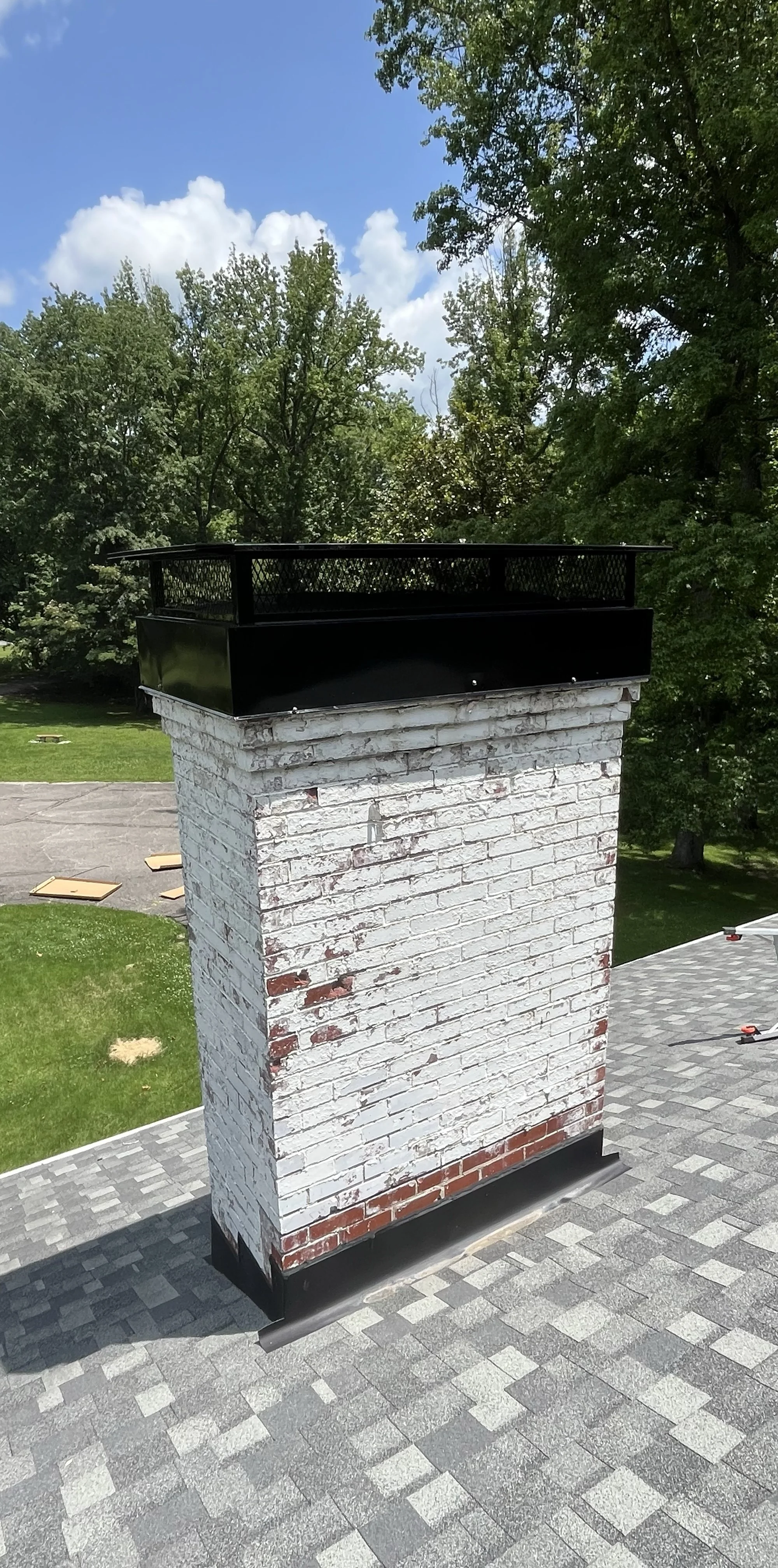 A brick chimney with a black metal cap on a rooftop, with a background of green trees, a partly cloudy blue sky, and a lawn.