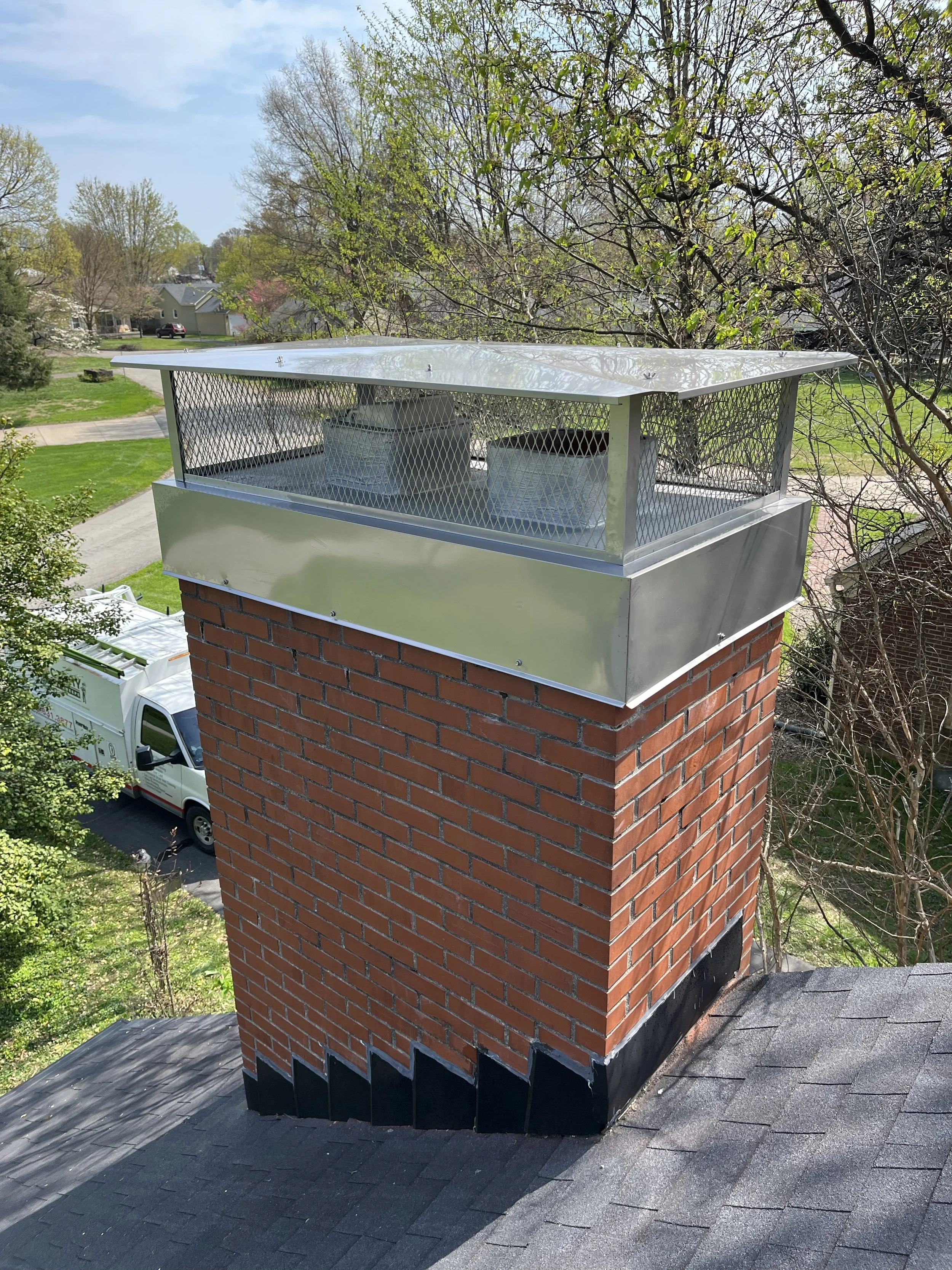 A brick chimney with a metal cap and a wire screen surrounding the top, situated on a shingled roof with a ridge vent. Trees and houses are visible in the background on a clear day.