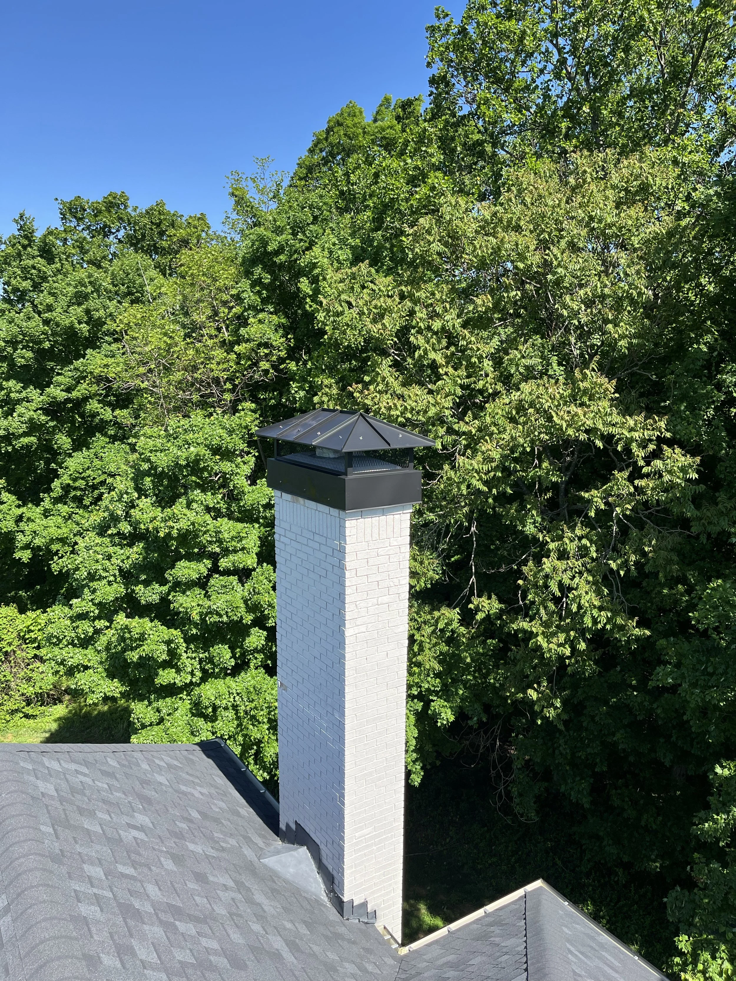 A white brick chimney with a black metal cap on a house roof, surrounded by green trees and clear blue sky.