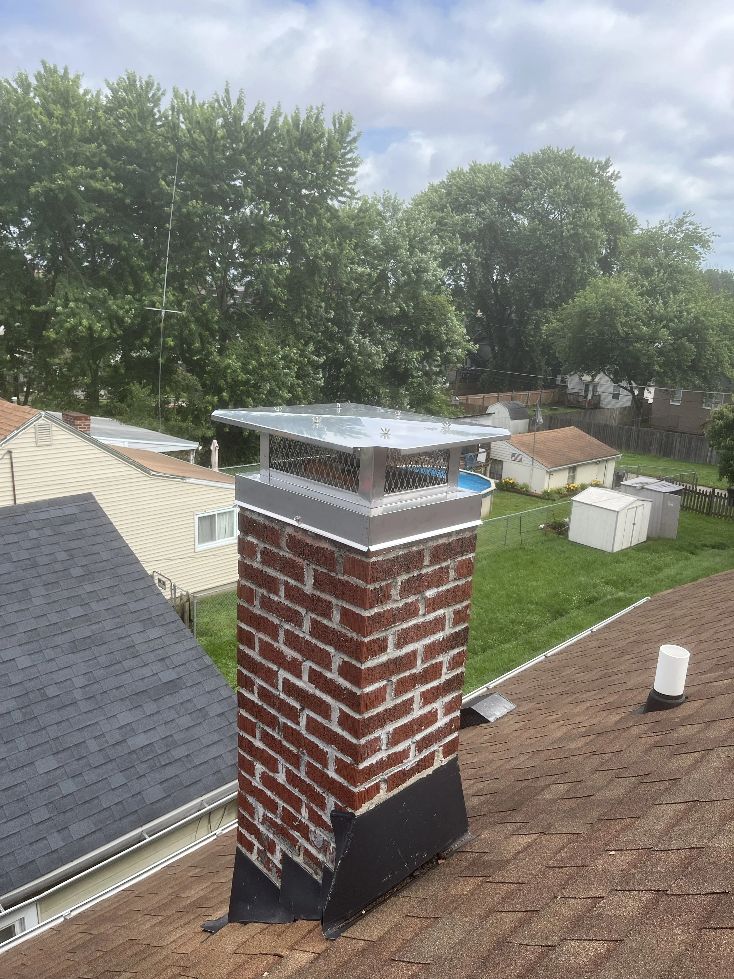 View of a house roof with a brick chimney topped with a metal cap; the roof is shingled in brown. Surrounding area includes trees, neighboring houses, and a backyard with a small shed. The sky is partly cloudy.