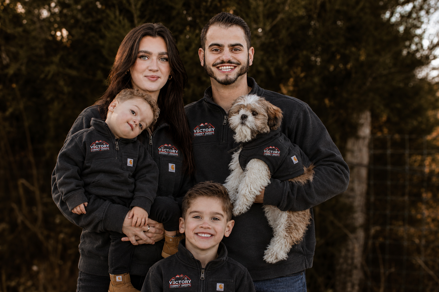 Family of four with two children and a dog outdoors during daytime, all wearing black jackets with logos, standing in front of trees.