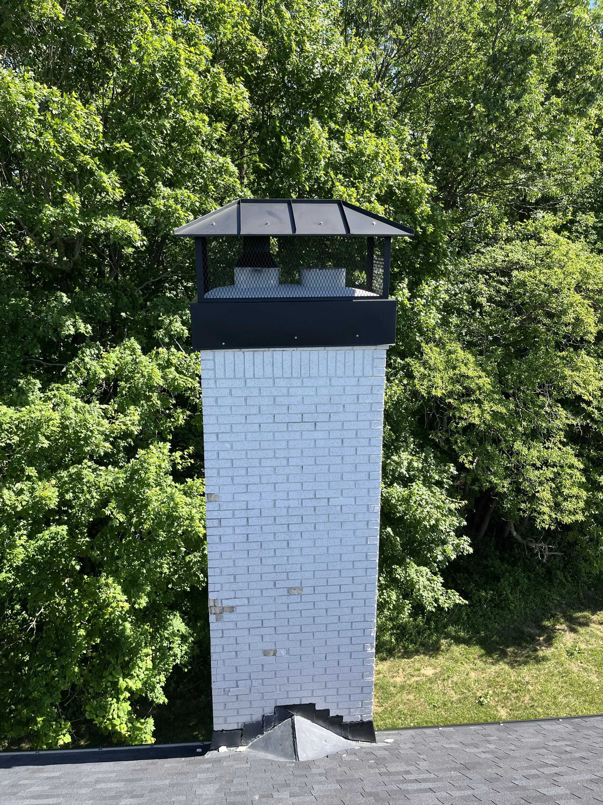 A white brick chimney on a black shingled roof with a metal cap, surrounded by green trees.