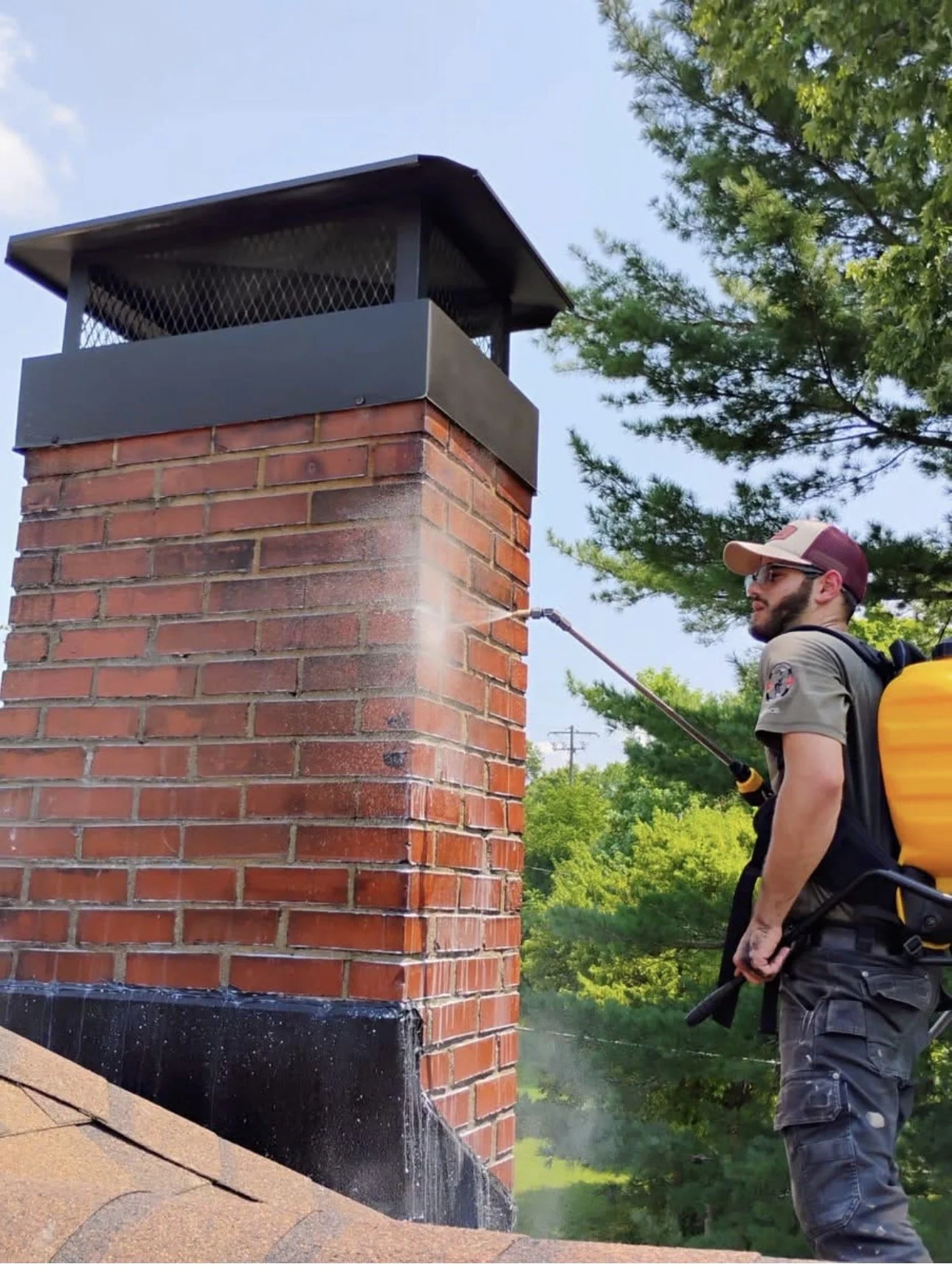 A man wearing a hat, sunglasses, and a uniform is spraying a chimney with a garden hose. He has a yellow backpack attached to the hose reel, which is dripping water on the brick chimney. There are trees and a blue sky in the background.