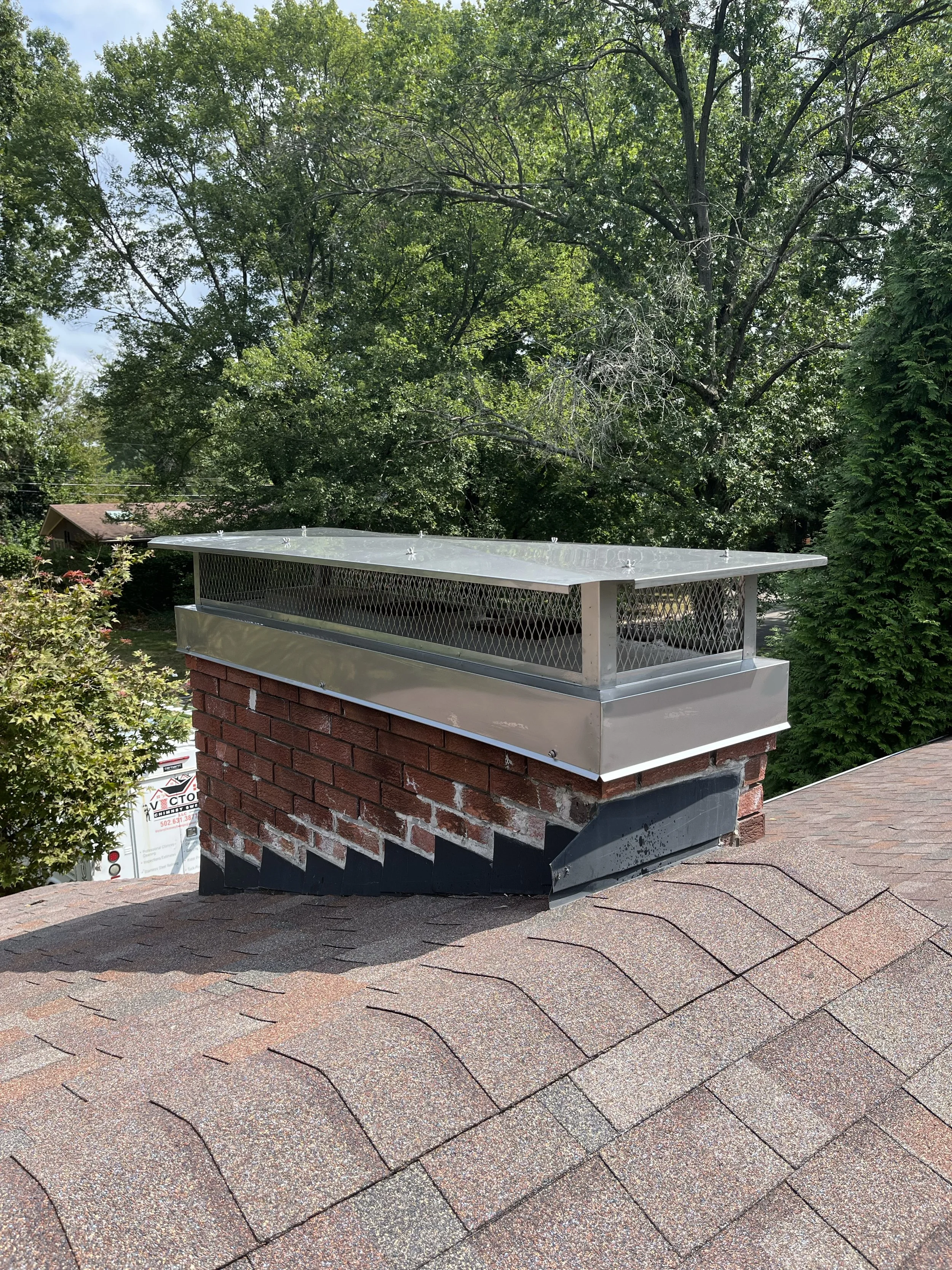 A brick chimney with a metal cap and guard on top of a sloped shingle roof, surrounded by green trees and plants.
