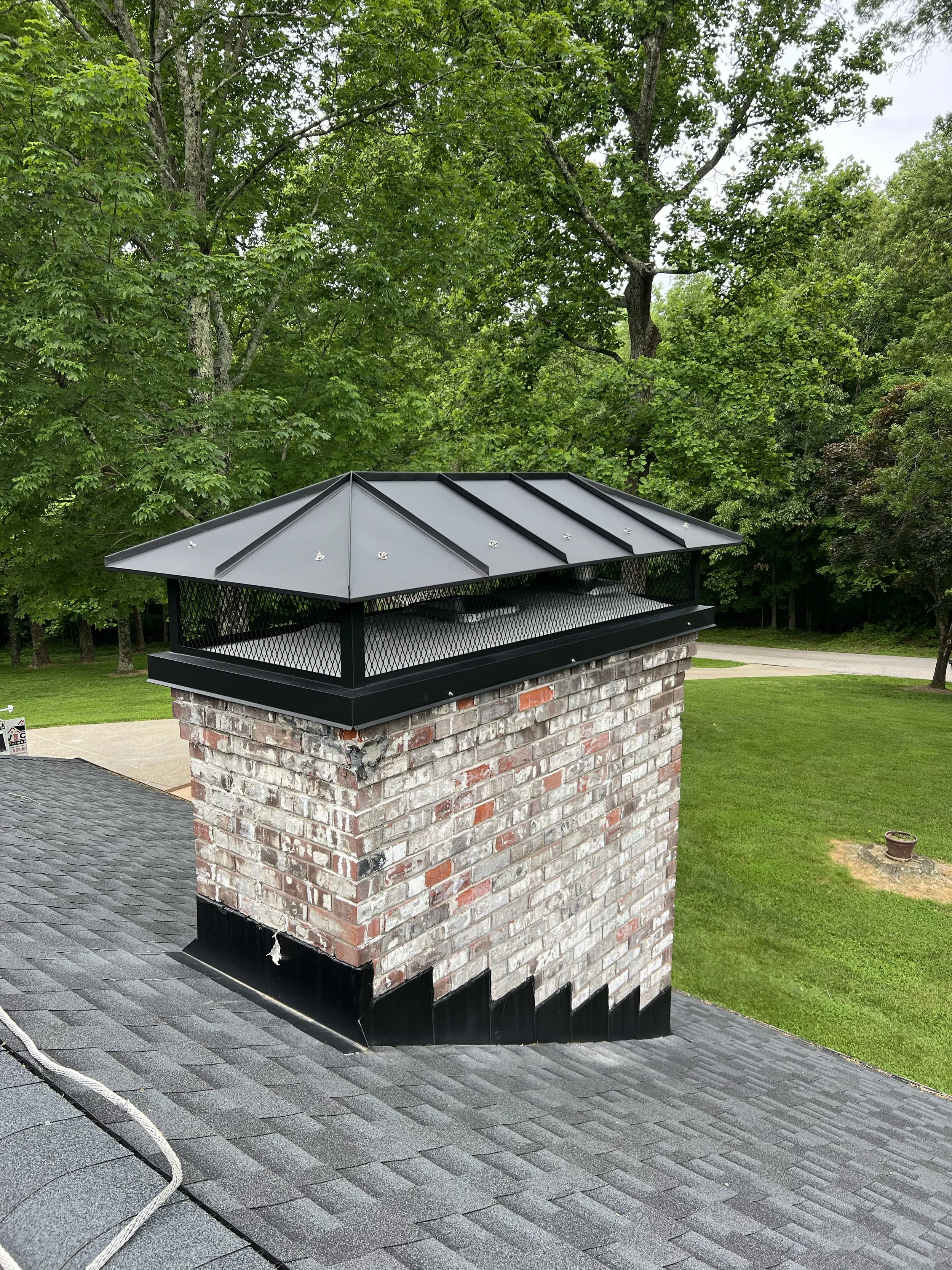 Close-up of a brick chimney with a metal cap, on a roof with gray shingles, surrounded by green trees and a lawn in the background.