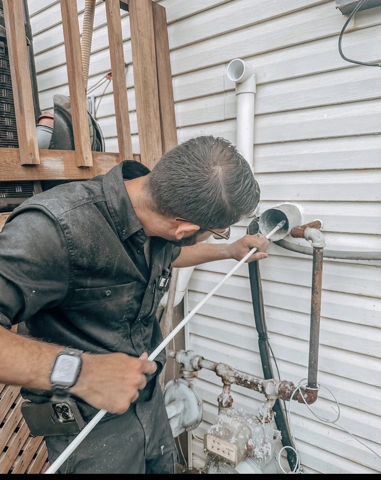 A man working on outdoor plumbing, repairing or installing pipes on the side of a house with white siding.