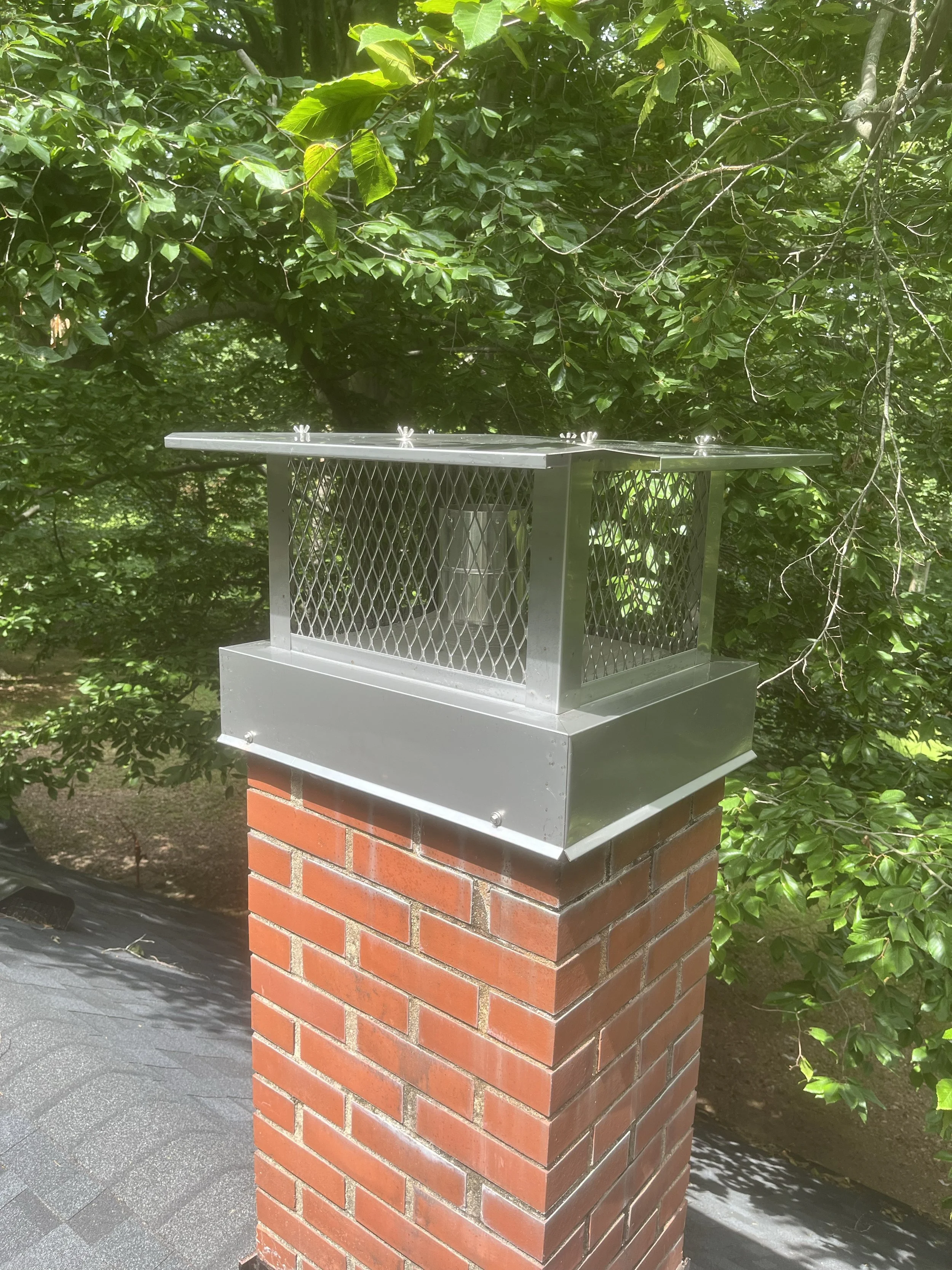 A metal bird feeder with a roof, mounted on a brick pillar, surrounded by lush green trees and bushes.